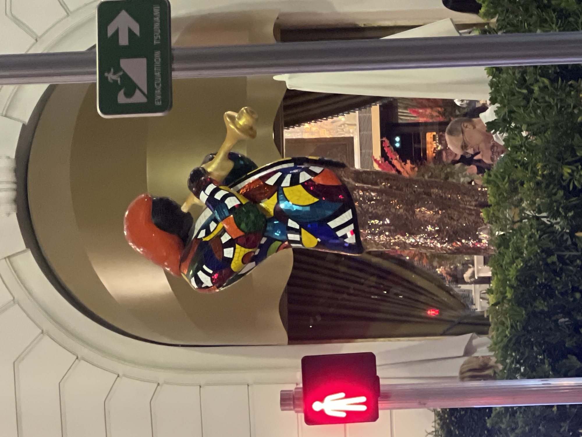 In Nice, France, a colorful, mosaic statue of a person playing a trumpet stands prominently near an arched entryway, possibly of a building. The statue is adorned with a red head covering and a vibrant, multi-colored jacket over a shimmering gold skirt. To the left, a pedestrian traffic signal displays a red illuminated walking figure, indicating it is not safe to cross. To the right, a green sign with a white pictogram of a person running from a wave and an arrow points to the right, indicating "EVACUATION TSUNAMI." In the foreground, lush green foliage partially obscures the lower portion of the scene, with several individuals partially visible behind the greenery, engaged in conversation.