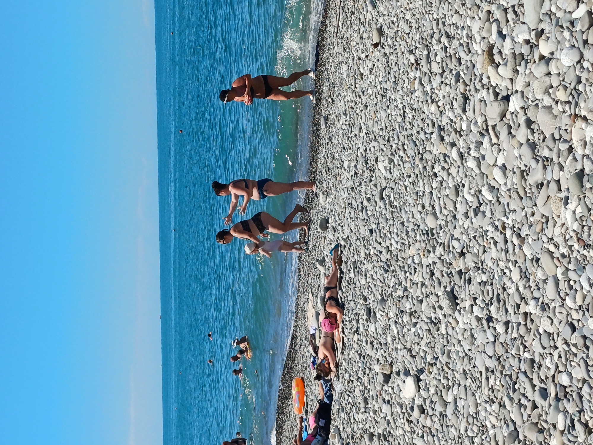 The image depicts a sunny day at a pebble beach on the coast of Sochi, Russia. In the foreground, several individuals are sunbathing on towels and mats scattered across the gray pebbles. Two women, one wearing a pink cap and the other with her hair in a bun, are lying down, while another person is partially visible on the left.
In the middle ground, near the water's edge, a group of people are standing and interacting. A woman in a patterned bikini is holding a small child, who is wearing a white sun hat. Beside them, another woman in a blue bikini appears to be talking to them. Further to the right, a woman in a black bikini and hat is standing near the water.
The background is dominated by the bright blue sea, with small waves gently lapping the shore. Several other people are visible swimming and playing in the water. The horizon line is clear, and the sky is a cloudless, vibrant blue, suggesting a warm and pleasant day. The scene captures a typical summer day at a popular Russian Black Sea resort.