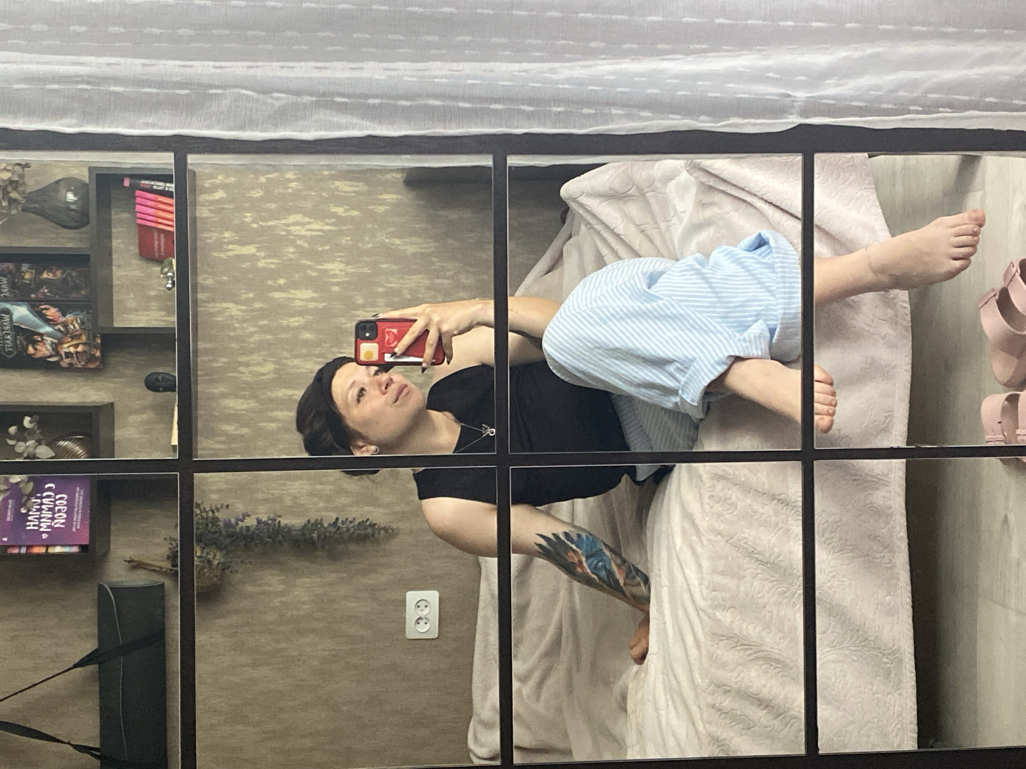 A young woman with a dark headscarf is sitting on a bed, taking a mirror selfie. She is wearing a black tank top and light blue and white striped pants. Her left arm, which has a tattoo, is resting on the bed. The bed is covered with a light pink comforter and a white patterned blanket. In the background, on shelves above the bed, are books, decorative items, and a vase. A white sheer curtain is visible to the right of the frame. The scene appears to be an indoor bedroom. The image is framed by dark grid lines, suggesting it is a reflection in a multi-paneled mirror.