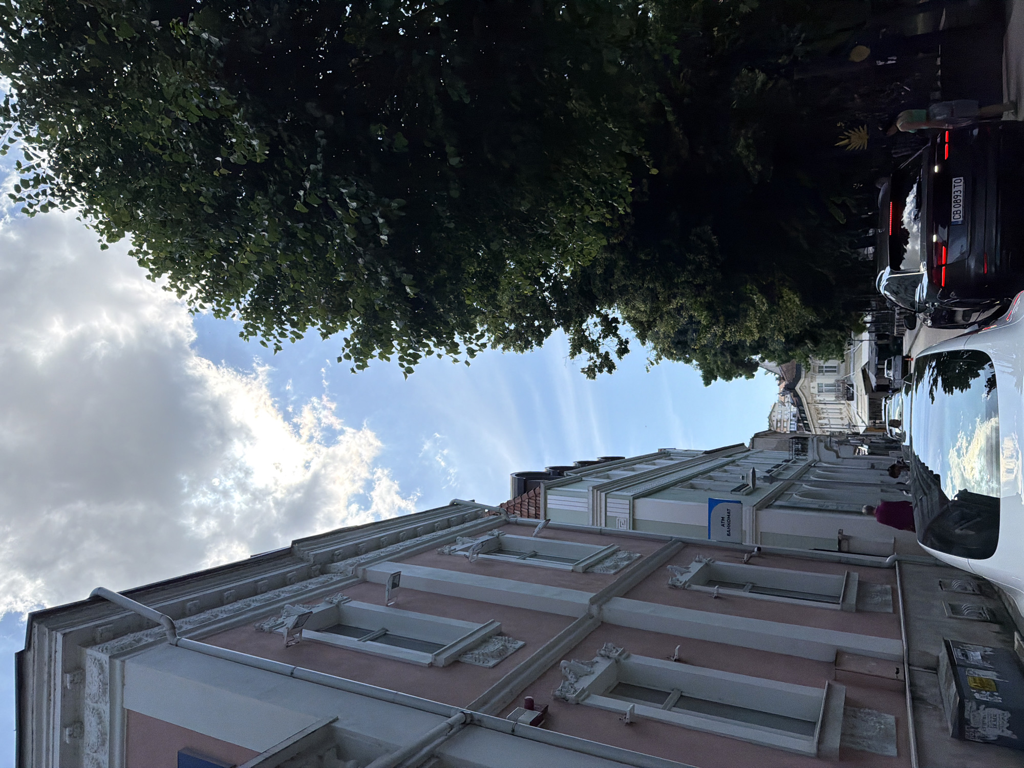 A street scene in a city, likely Pleven, Bulgaria, captured during the daytime with clear blue sky and scattered white clouds. The perspective is tilted, looking upwards along a street lined with buildings and lush trees.
The upper portion of the image is dominated by the dense green foliage of trees, providing a canopy over the street. Sunlight filters through the leaves and illuminates parts of the sky.
On the right side of the image, ornate buildings line the street. The closest building features a pastel pink facade with intricate white trim, including decorative elements around its windows and a detailed cornice. Drainpipes are visible running along the building's exterior. Further down, other buildings with similar classical architectural styles and lighter hues (greenish-grey) can be seen.
Along the street, several cars are parked. A dark-colored SUV is prominently visible, displaying a Bulgarian license plate "CB 0893 TO". The reflection of the sky and clouds is visible on its surface. Part of a white car is also visible in the immediate foreground.
People are present on the sidewalk. One individual wearing a purple top is walking near a building that has a blue sign with white text, which reads "ATM БАНКОМАТ" (ATM Bankomat). Another person in light green shorts is visible further down the street near the parked vehicles. The scene depicts a typical urban environment with a mix of historical architecture, greenery, and daily street activity.