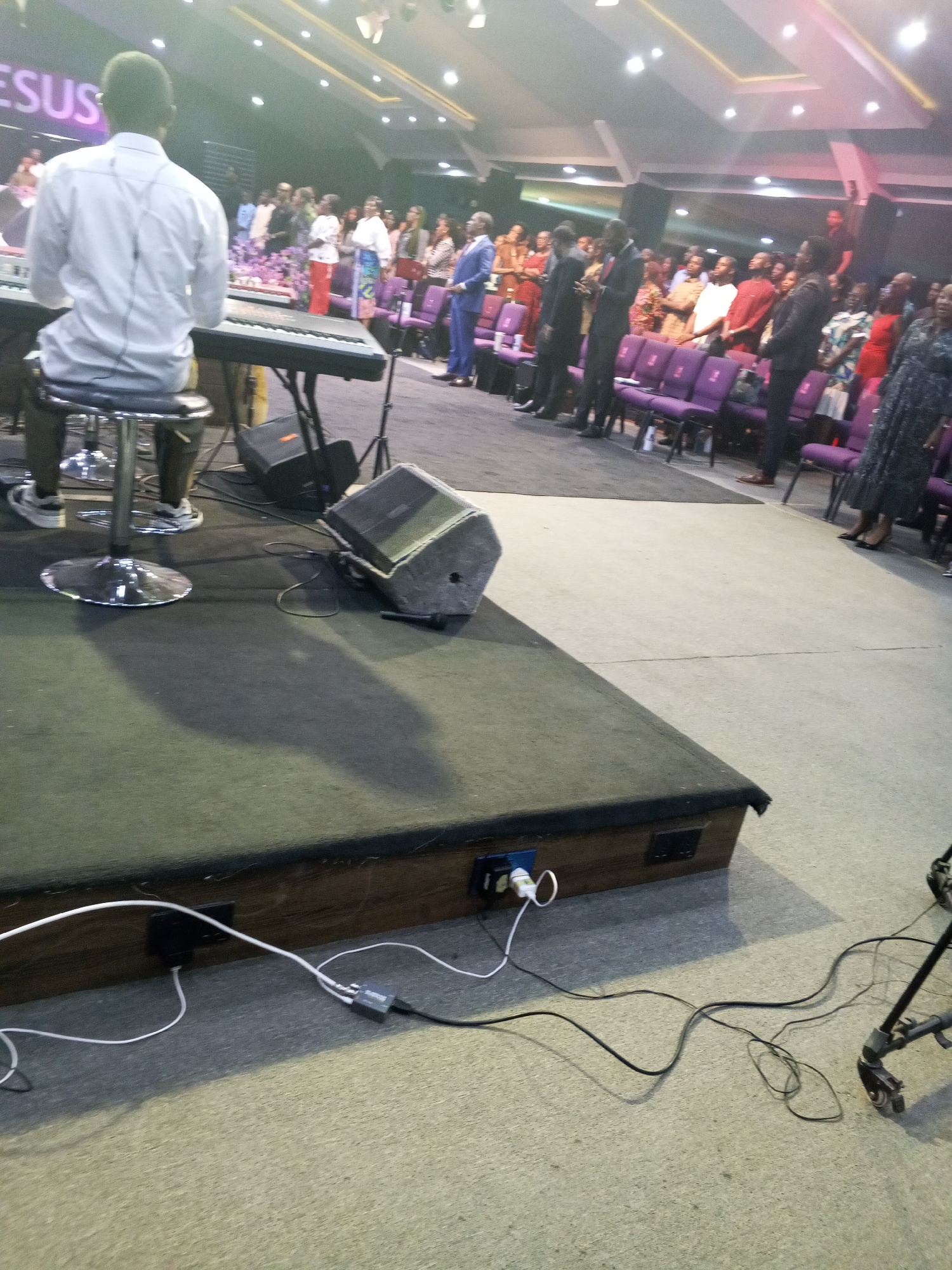 The image depicts a scene inside a church in Abuja, Nigeria. A man in a white shirt is seated at a keyboard on a stage. In front of him are rows of people standing and facing forward, presumably during a service. To the right, there are several rows of purple chairs behind the people. The stage itself is carpeted and has visible electrical outlets and cables running along its edge. A large loudspeaker is placed to the right of the keyboard. The lighting is bright, with ceiling lights illuminating the space. The word "JESUS" is visible in the upper left corner, suggesting the religious nature of the event. The setting appears to be a large auditorium or sanctuary.