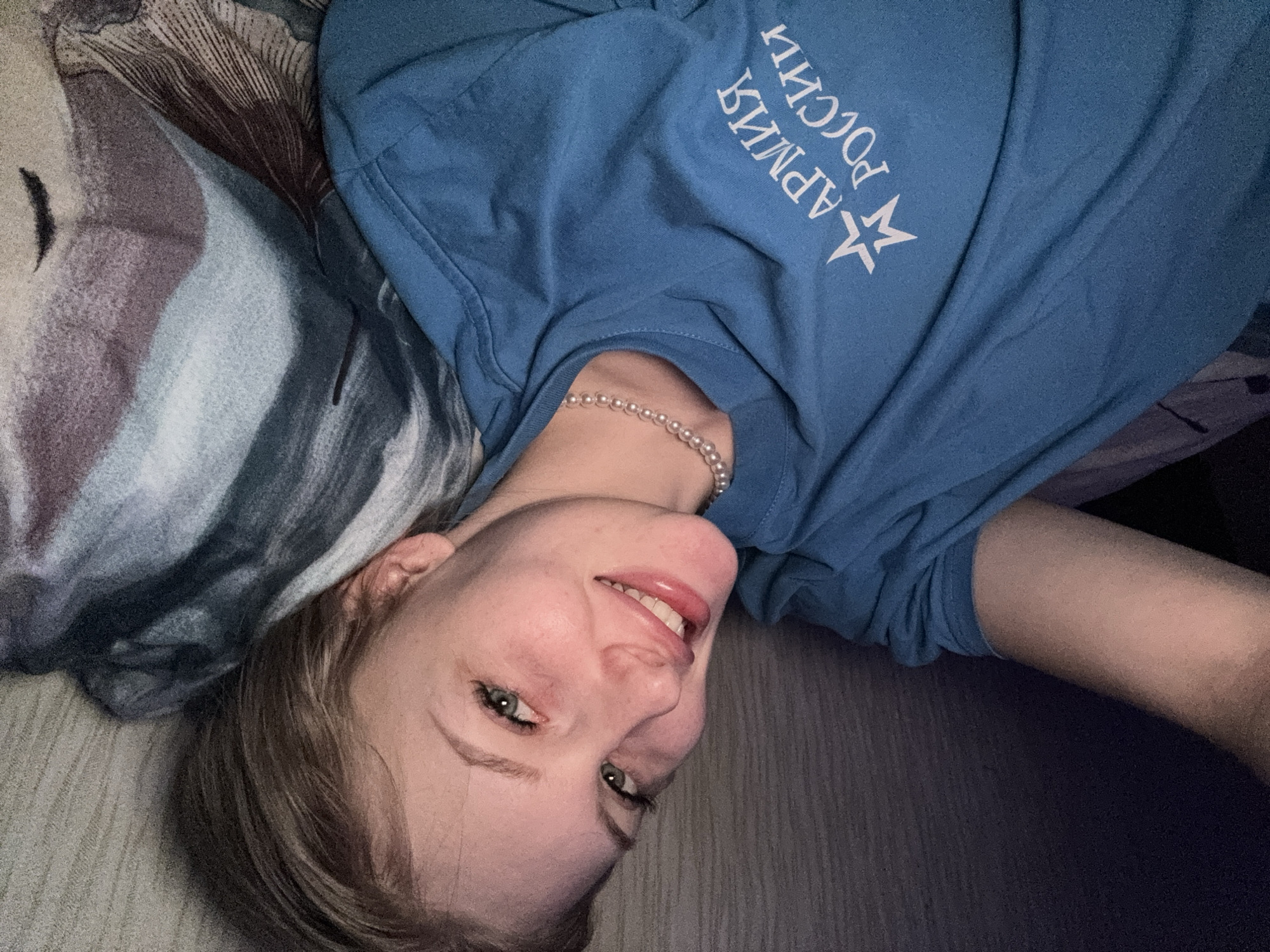 An indoor, close-up, eye-level shot captures a young woman lying down and looking directly at the camera with a cheerful, playful expression. Her head is tilted to the right, and she is smiling, showing her teeth and a bit of her tongue. She has light brown hair, green eyes, and fair skin.
The woman is wearing a light blue short-sleeved t-shirt with white text and a star symbol on the front. The text, in Cyrillic, reads "АРМИЯ РОССИИ" (Army of Russia). Around her neck, she wears a pearl necklace or choker. She is resting her head on a patterned pillow or blanket with abstract designs in shades of grey, blue, purple, and brown. The background on the right and bottom shows a light-colored surface, possibly a floor or bed. The lighting is soft and even, suggesting an indoor setting in Кондратово, Russia.