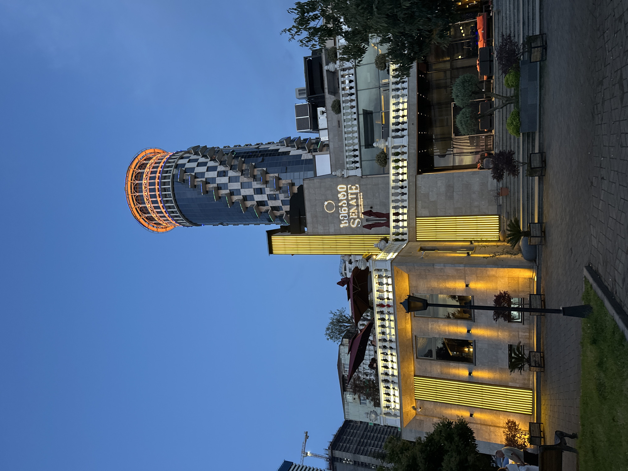 The image shows the illuminated exterior of the "Batumi Senate" restaurant and adjacent building in Batumi, Georgia, at dusk. The cylindrical building has a checkered facade and an orange illuminated top. The restaurant's sign "Batumi Senate" is visible. The building is illuminated with linear yellow lights. A portion of the pedestrian walkway is visible.