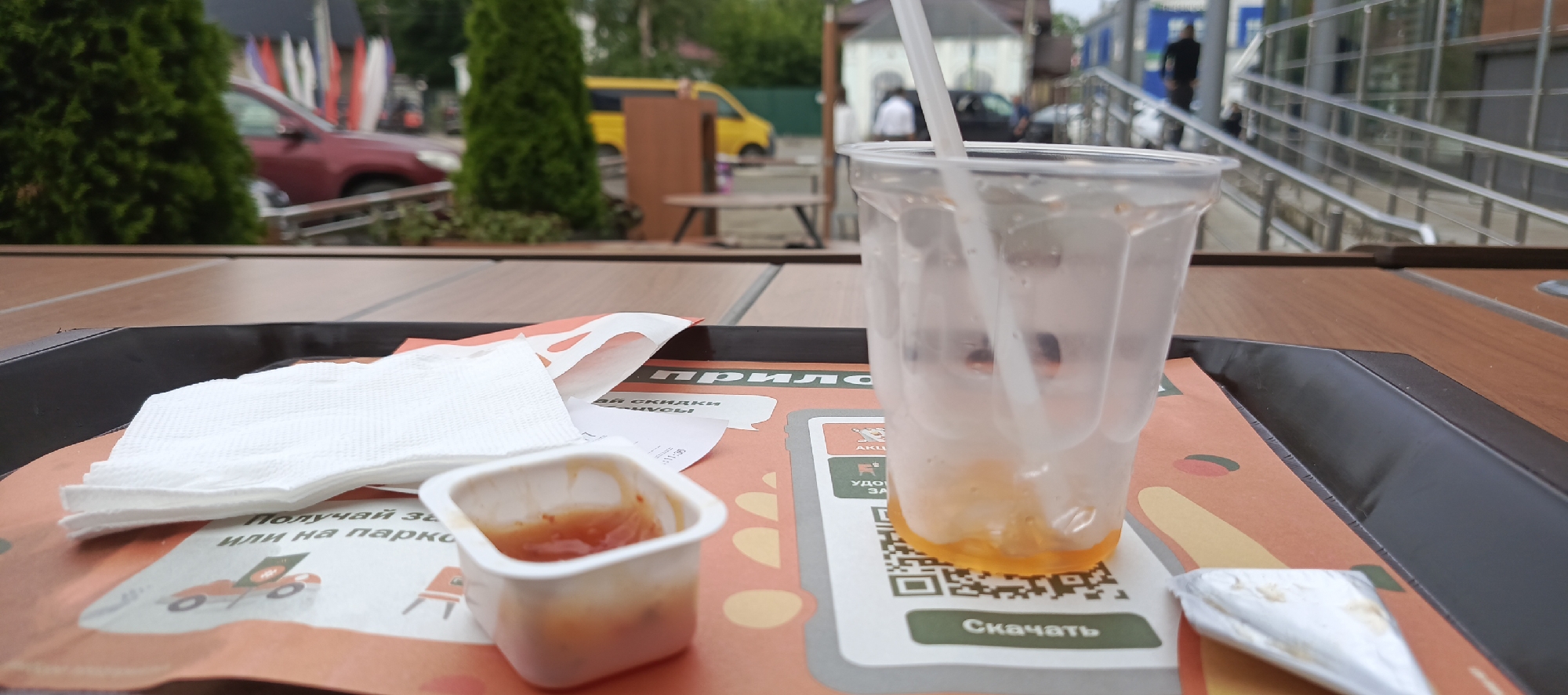 On a wooden table, a dark tray holds the remains of a meal. In the foreground, a white napkin is folded neatly, next to a small white container with a reddish sauce, likely ketchup. To the right, a clear plastic cup, partially filled with a yellow or orange liquid and an empty straw, sits on top of a printed mat. The mat has a graphic design with various shapes and text. A QR code is visible on the mat, below the cup. Partially obscured by the cup and napkin is a receipt. In the background, a red car is parked near some green trees. Further in the distance, a yellow van is visible, along with some buildings and railings. People are faintly visible in the background, suggesting an outdoor dining area. The scene appears to be at a fast-food restaurant or a similar establishment. The lighting suggests it is daytime, possibly overcast. The text on the mat includes "Скачать" (Download) and other Cyrillic characters. The receipt also contains Cyrillic text, though it is difficult to read clearly.