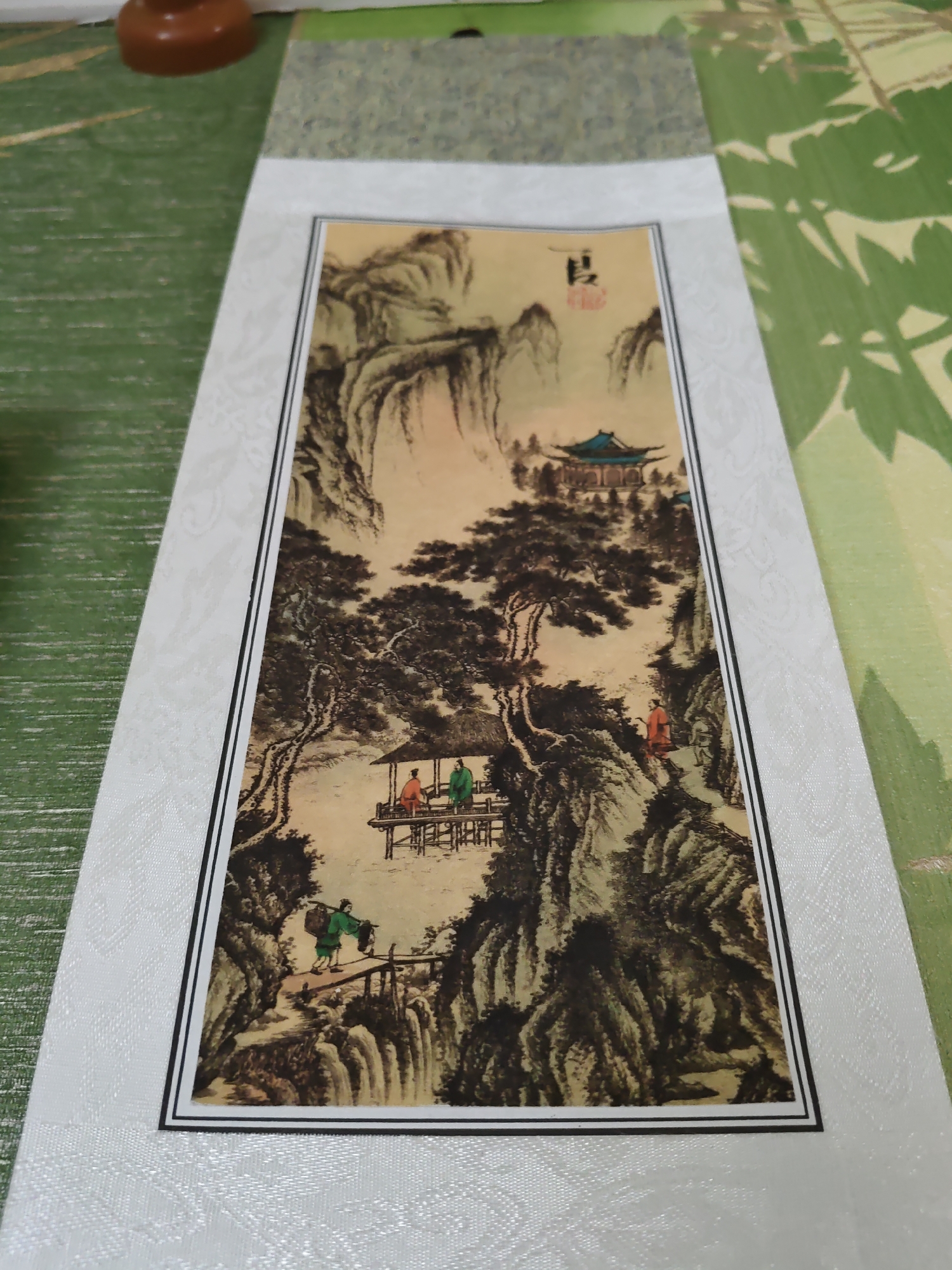 The image shows a scroll painting of a traditional Chinese landscape. The painting depicts a mountainous scene with a pavilion and figures. A person in a green robe is walking across a bridge, carrying a basket on their shoulder. Further up the path, another person in an orange robe is walking. In the pavilion, two figures are seated. A large pine tree is prominently featured in the foreground. In the upper right corner of the painting, there are Chinese characters and a red seal. The scroll is mounted on a textured white border and is placed on a green surface with a subtle pattern. The painting is illuminated by natural light. There is no information in the image that can be used to determine its location in Rostov-on-Don, Russia.