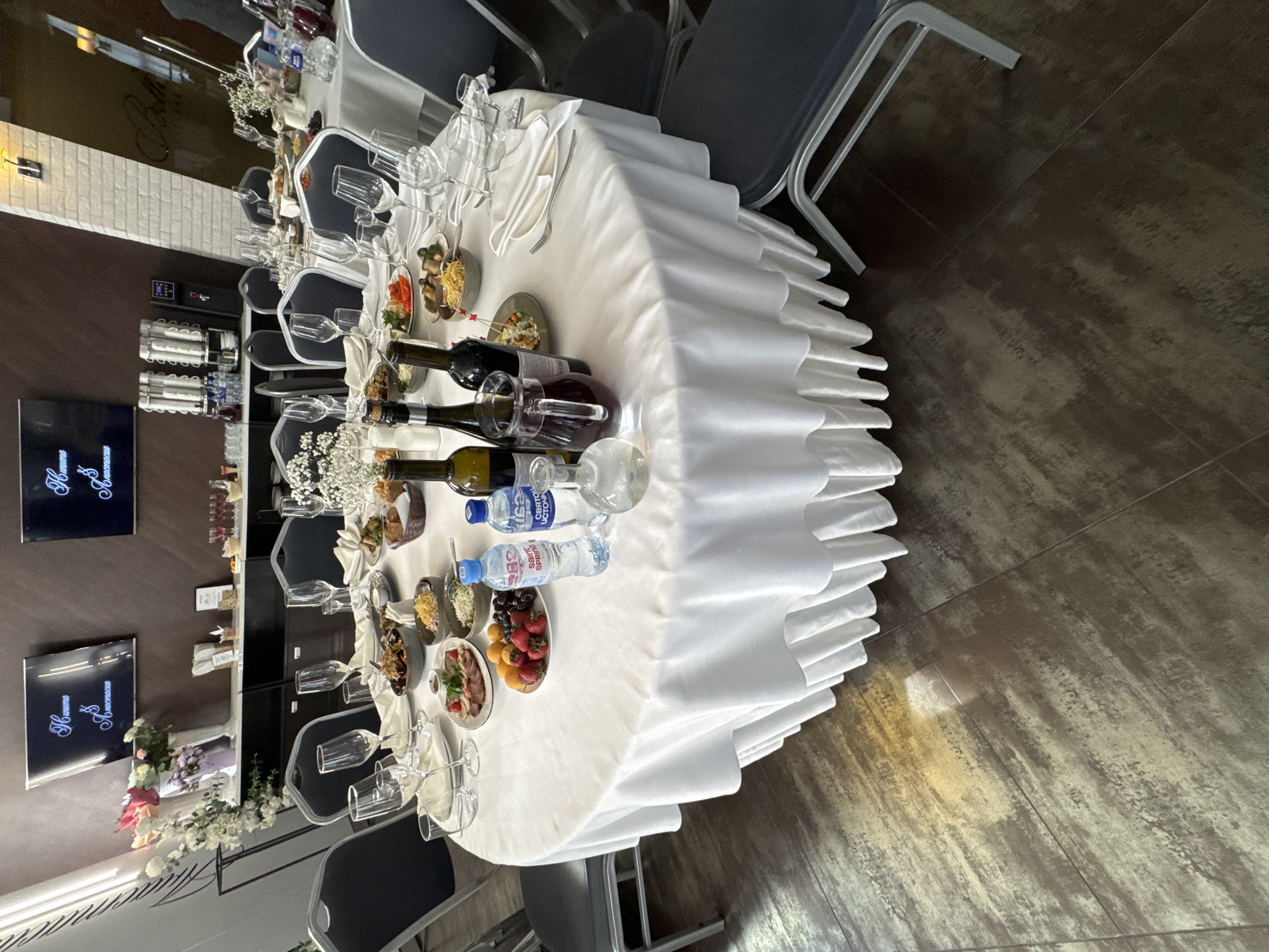 The image shows a round banquet table set for a formal meal, likely a wedding reception given the screens displaying elegant script. The table is covered with a white tablecloth that drapes to the floor with a ruffled edge. On the table are numerous place settings, each with a white napkin, silverware, and glassware. Several plates of food, including what appears to be meat dishes, salads, and fruit, are arranged around the table. Two bottles of wine and a pitcher of dark liquid, possibly sangria or another mixed drink, are prominently featured. Two bottles of Saint Spring mineral water are also present. In the background, there are chairs around other tables, suggesting a larger event. The room appears to be a banquet hall or restaurant with dark flooring and brown walls. Two large flat-screen televisions are mounted on the wall, displaying text. There are also some decorative elements, including a vase of flowers and some small floral arrangements on the table. The scene is well-lit, with a bright overhead light reflected on the floor. There are no people visible in the image. The image does not provide any specific information to identify the location as Irkutsk, Russia.