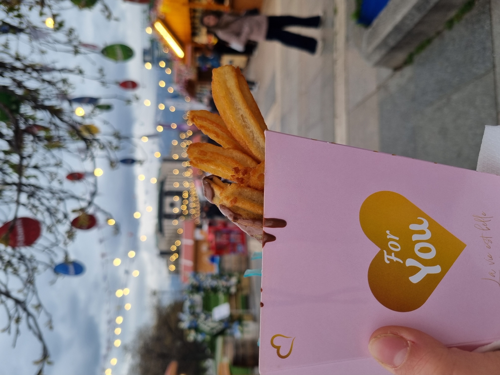 The photograph shows a close-up of churros in a pink paper container with a golden heart and the words "For You" printed on it. Some of the churros are coated in chocolate. The container is being held by a hand. In the background, there's a blurred view of a Christmas market with lights and decorations. In the blurred background, there are also bare tree branches with hanging colorful ornaments, and a person is walking in the distance. Based on the decorations and the lights, it appears to be taken during a festive season. The photo was taken at the Christmas market in Sofia, Bulgaria.
