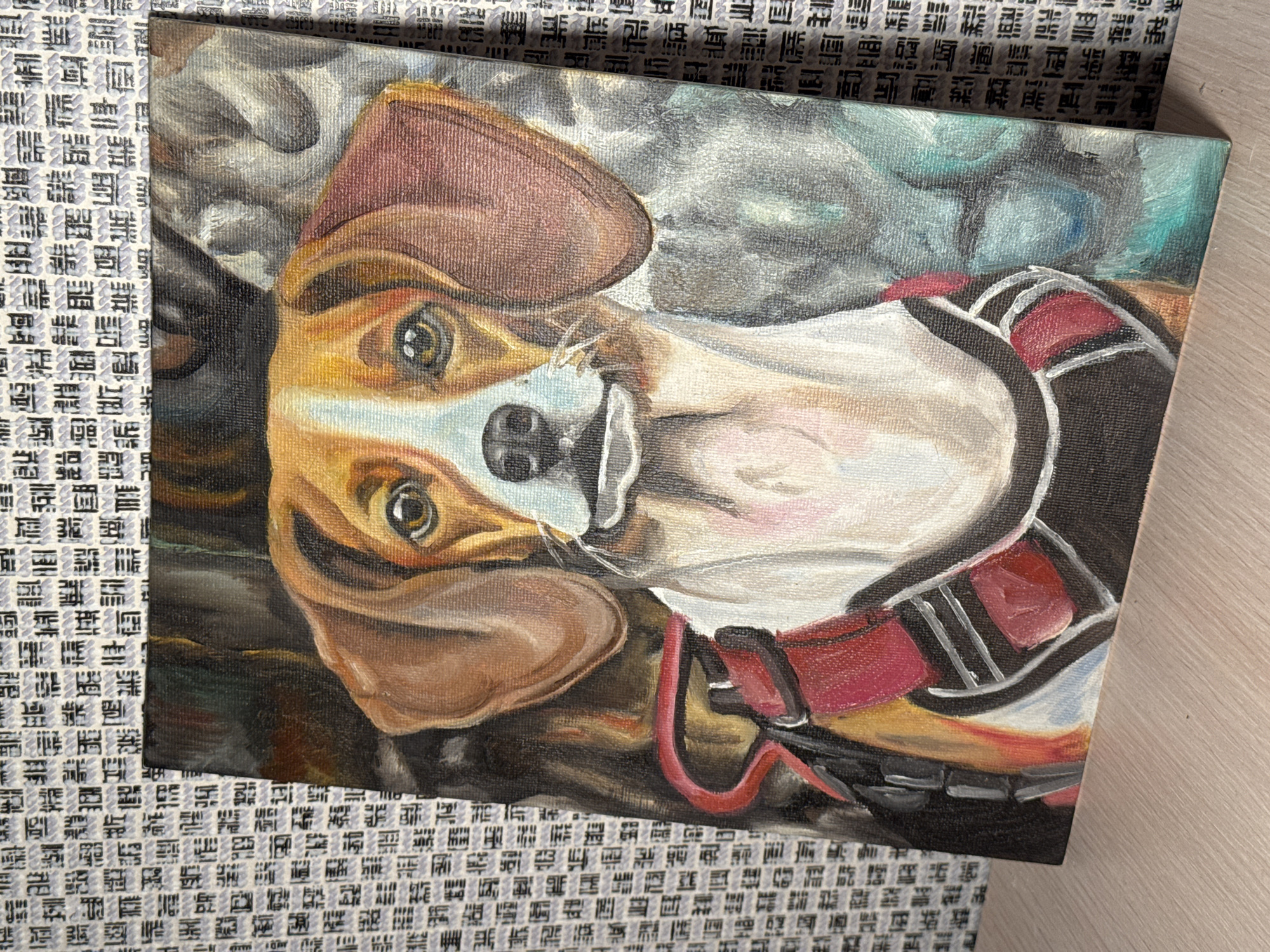 The image shows an oil painting of a brown and white dog wearing a red and black harness. The background is a mix of grey, blue, and brown hues. The painting is placed on a surface with a textured white and grey pattern next to a light brown surface.