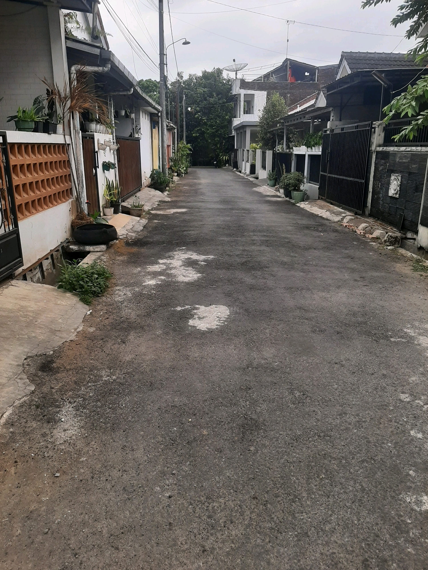 The image shows a narrow, asphalt-paved street in a residential area. The street is lined with houses on both sides, with a mix of architectural styles. Some houses have white walls and wooden gates, while others feature darker, more modern designs with black metal fences. Potted plants are visible along the sidewalks and in front of some houses, suggesting a well-maintained but somewhat cluttered residential environment. The sky is overcast, indicating a cloudy day. The overall impression is that of a quiet, possibly working-class neighborhood street. This scene is located in the city of Sumampir, Indonesia.