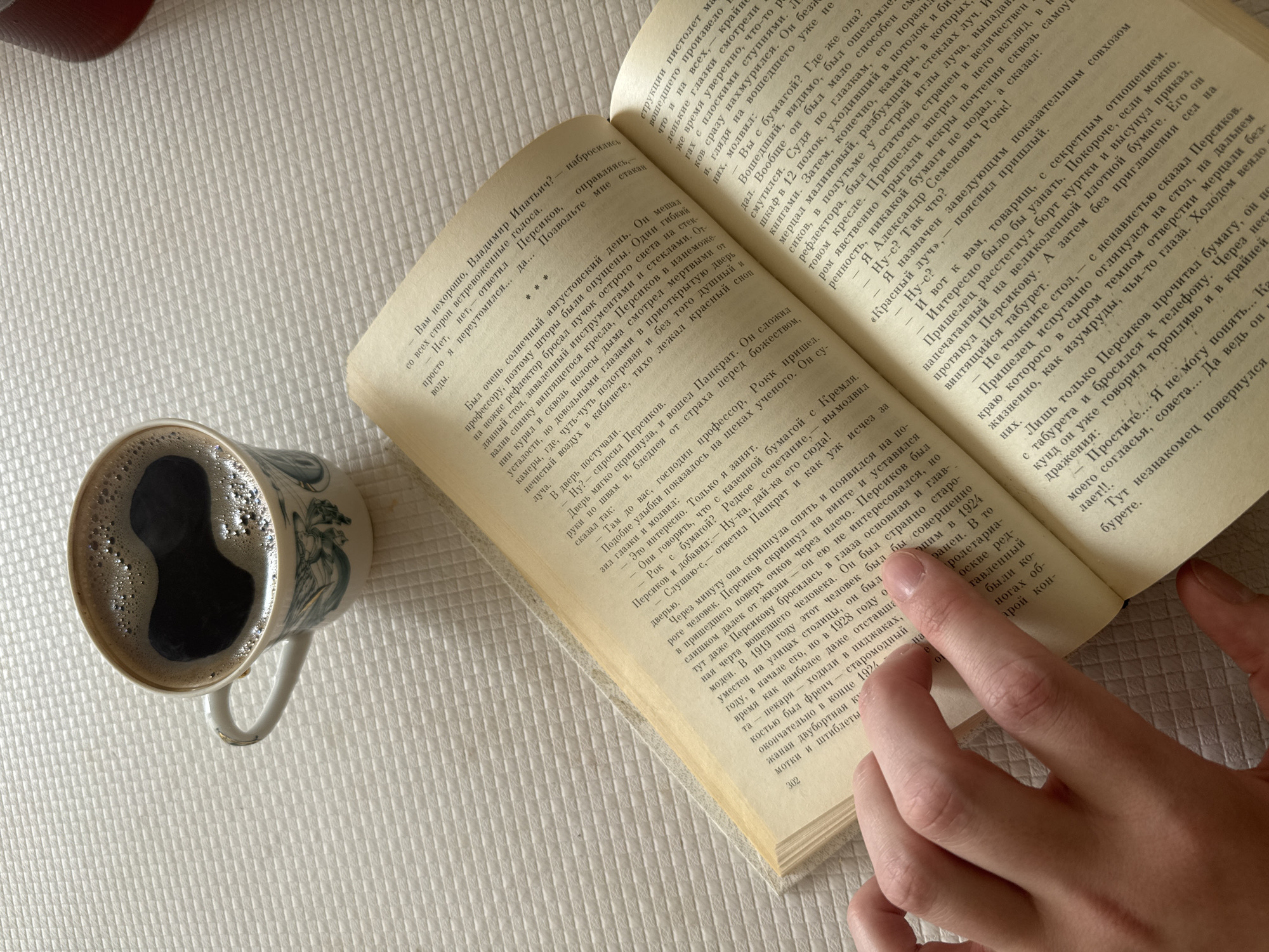 The image displays an intimate indoor scene featuring an open book, a mug of coffee, and a person's hand, all resting on a white, textured surface. The primary activity depicted is reading, with a finger gently holding open the book's yellowed pages.
The book is a physical copy with extensive text in Russian. Several paragraphs and dialogue snippets are visible. Phrases such as "Вам нехорошо, Владимир Ипатьич?", "Персиков", "стакан воды", "Был очень солнечный августовский день", "профессору", "Рок пришел", "Александр Семенович Рокк!", and "Простите... Я не могу понять..." suggest a narrative from a literary work, possibly a novel. The page number "302" is visible at the bottom of the right page.
To the left of the book, a ceramic mug with a subtle, possibly blue-and-white, decorative pattern holds a dark beverage, likely coffee or espresso, topped with a layer of foam. The white textured surface, resembling a waffle-weave fabric, serves as a clean and bright background. The lighting appears soft and indirect, contributing to a calm and contemplative atmosphere. The image evokes a sense of relaxation and intellectual engagement, typical of a quiet moment spent reading and enjoying a warm drink. No specific location cues are discernible beyond an indoor setting.