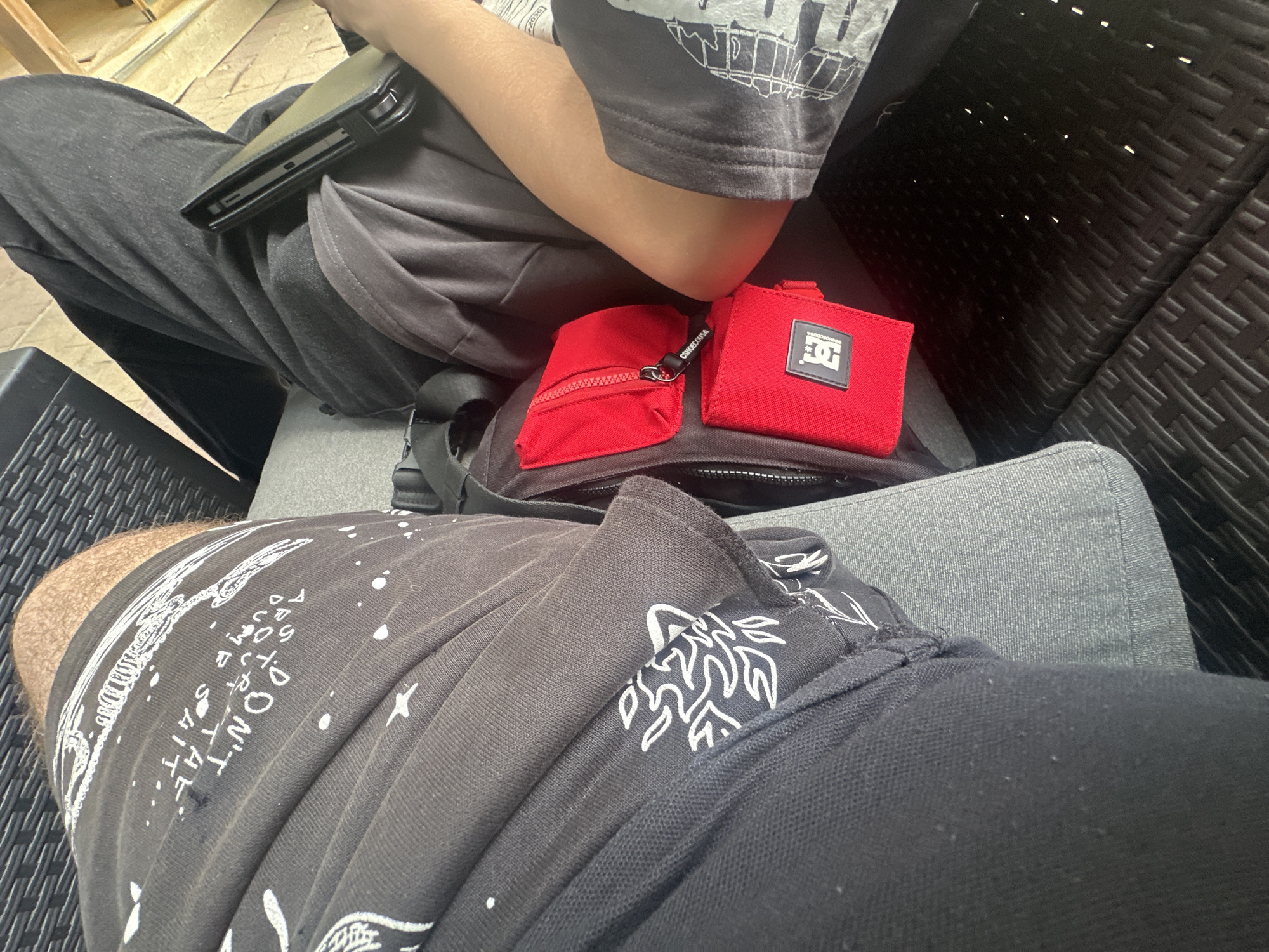 This image, captured in the city of Lom, Bulgaria, presents a high-angle view of two individuals seated in an outdoor or semi-outdoor environment. The person on the left displays their upper right thigh and lower torso, clad in dark grey shorts or sweatpants featuring white graphics, including the partially visible phrase "DON'T FEAR" and various abstract patterns. A section of a hairy thigh is visible at the top. This individual is positioned on a light grey, fabric-covered cushion. The person on the right is shown with their lower torso, right forearm, and elbow, along with their upper thigh. They wear dark grey pants and a dark grey t-shirt with a graphic on the lower right that includes grid-like elements and text. A dark-colored protective case for an electronic device is held horizontally across their lap. This individual is seated on a dark brown/black chair or bench characterized by a woven wicker pattern. Situated between the two individuals is a black utility bag or fanny pack, to which two red accessory pouches are attached. The upper red pouch is zippered, with a white 'CSHOE.USA' inscribed on its zipper pull. The lower red pouch displays a black patch featuring a white 'DC' logo. The area behind the right person reveals a light-colored surface, possibly flooring. The scene is illuminated consistent with daytime conditions.