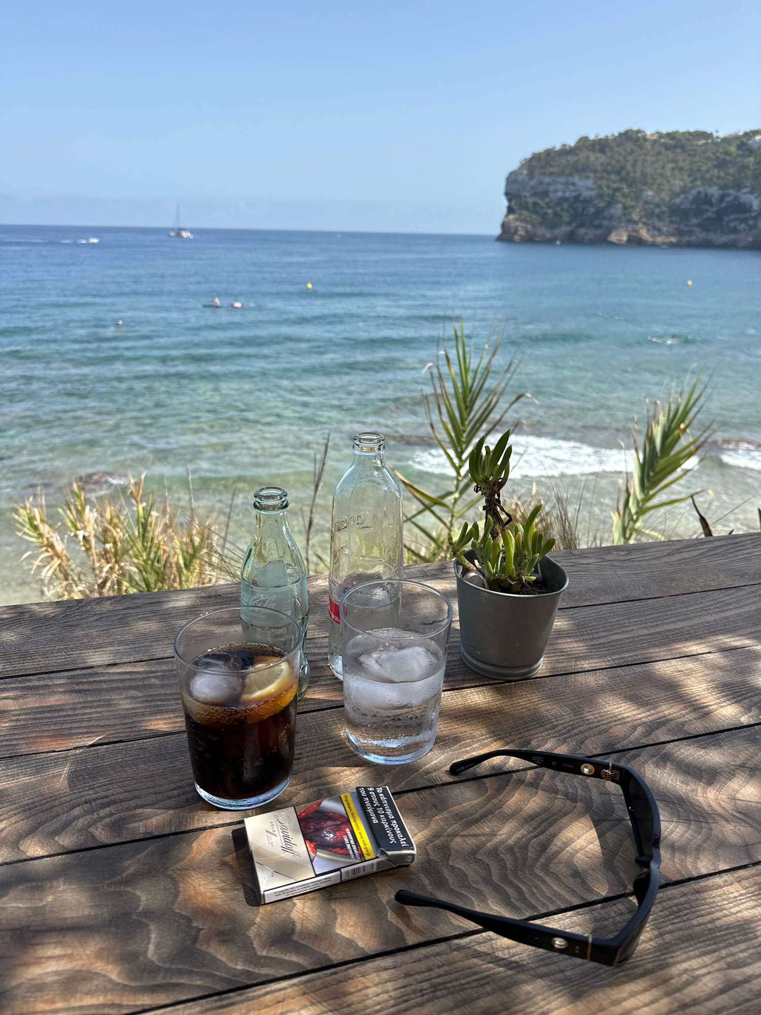 A table outdoors at a beachside cafe in Xàbia/Jávea, Spain, displays refreshments and accessories against a backdrop of the Mediterranean Sea. On the wooden table, a glass of dark liquid with ice and a lemon slice sits next to a glass of water with ice. Two glass bottles, one clear and one greenish, stand behind the glasses. A small potted succulent plant is positioned to the right. In the foreground, a pack of cigarettes and a pair of black sunglasses are laid out. The sea is visible in the background, with a few sailboats on the horizon and a rocky, tree-covered cliff to the right. The sky is clear and blue.