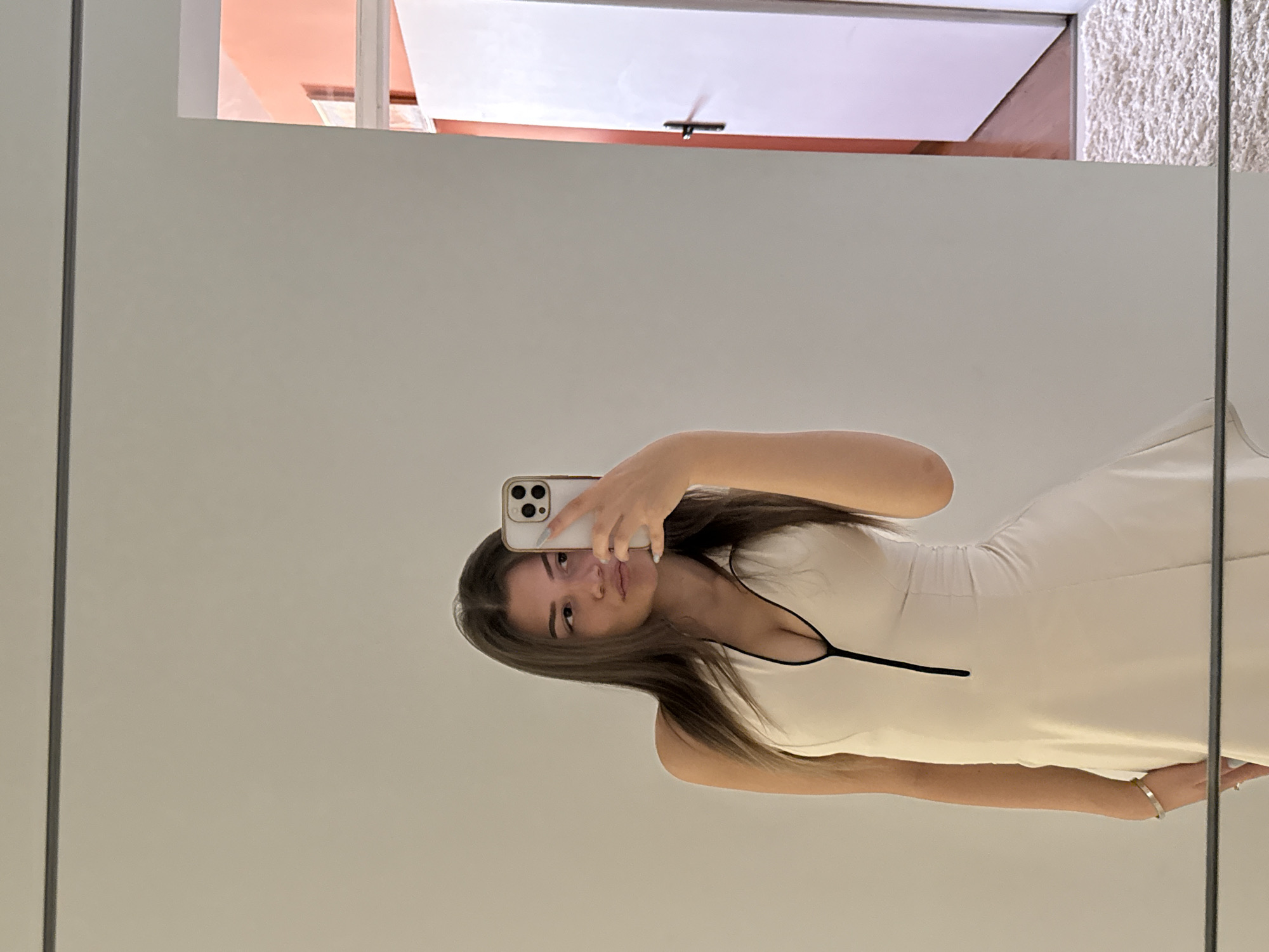 A young woman is taking a mirror selfie in a room. She is wearing a light-colored halter dress with a black zipper detail down the front. She has long, dark hair and is looking at her phone, which she is holding up to take the picture. Her left arm is bent, and she is wearing a bracelet and a ring. The background is a plain, light-colored wall, with a doorway and a sliver of a room with coral-colored walls visible to the right. A white rug is partially visible on the floor. The image appears to be taken indoors, likely in a residential setting, and does not provide any specific indicators to place it in Sofia, Bulgaria.