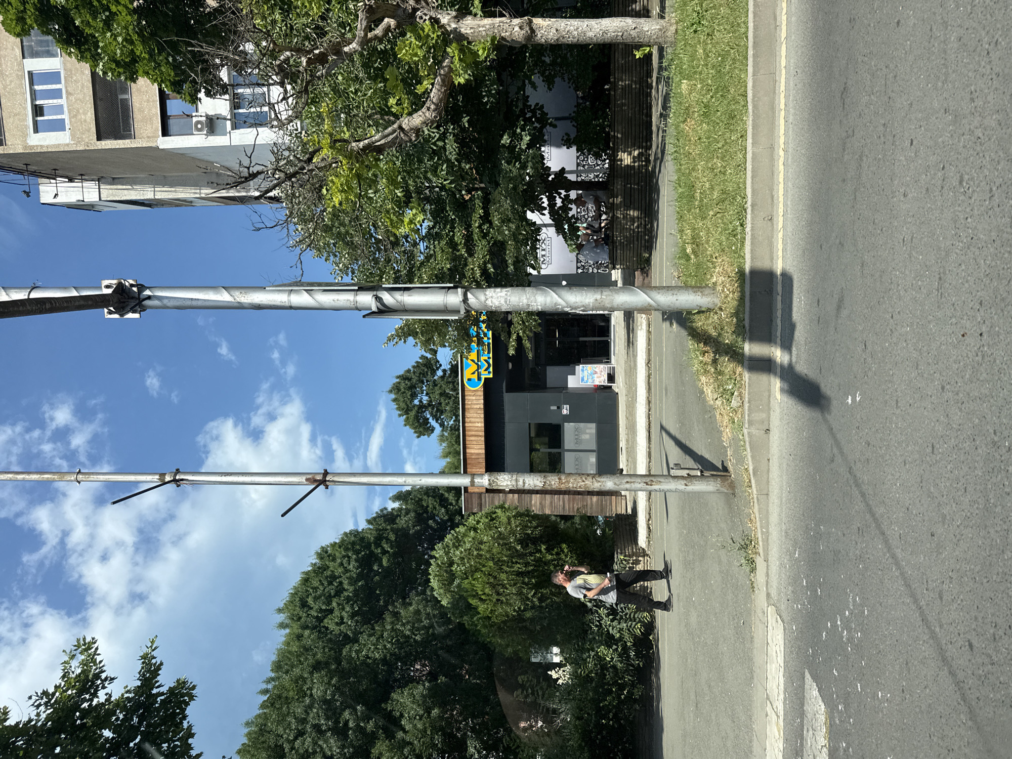 A full shot captures a street scene with a modern building on the left, characterized by several windows and an air conditioning unit. Adjacent to it, a dense canopy of green trees offers shade. In the foreground, a metal pole stands vertically, supporting what appears to be traffic or utility equipment. A person is walking on the sidewalk, wearing a t-shirt and dark pants, engrossed in a phone call. Behind them, a contemporary building houses a "MIX Market" with visible signage. The sky is clear and blue, suggesting a sunny day, likely during daylight hours. The pavement is made of asphalt, with a grassy area and a curb separating it from the sidewalk. Shadows cast by the trees and a pole indicate the sun's position. The overall scene depicts a typical urban street environment.