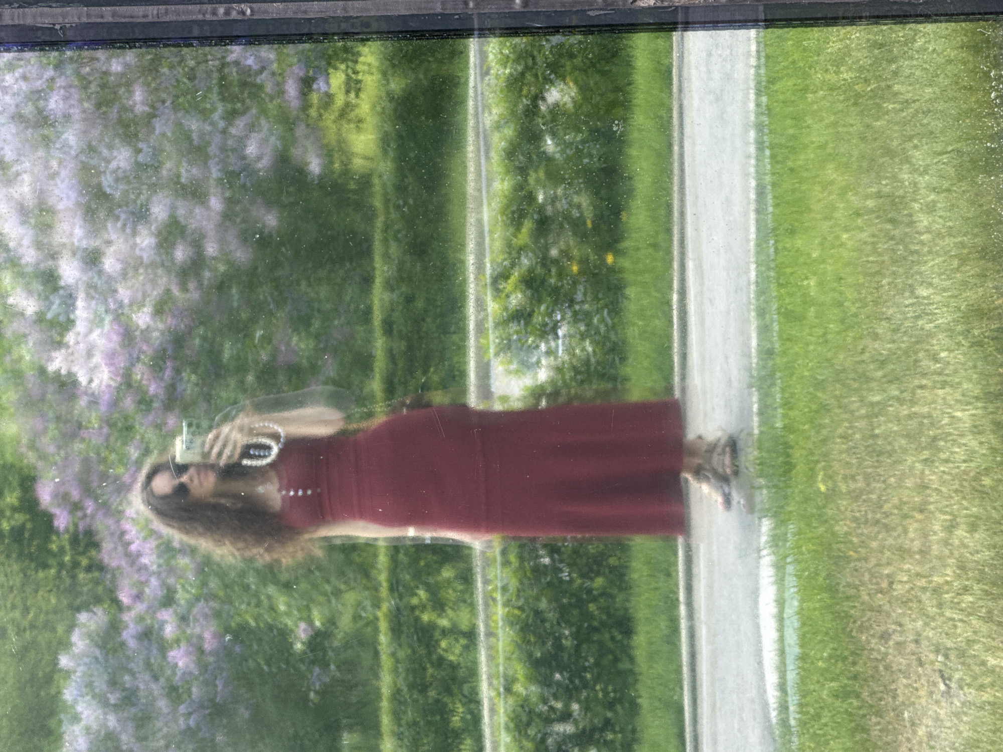 The image shows a reflection of a woman taking a photo of herself in a window. The woman has long brown hair and is wearing sunglasses and a long red dress. She is holding a camera in her hands and has a bracelet on her wrist. The reflection also shows trees, flowers, and a path. The flowers are lilac, a common ornamental plant in Moscow, Russia. The scene appears to be outdoors, possibly a park or garden setting.