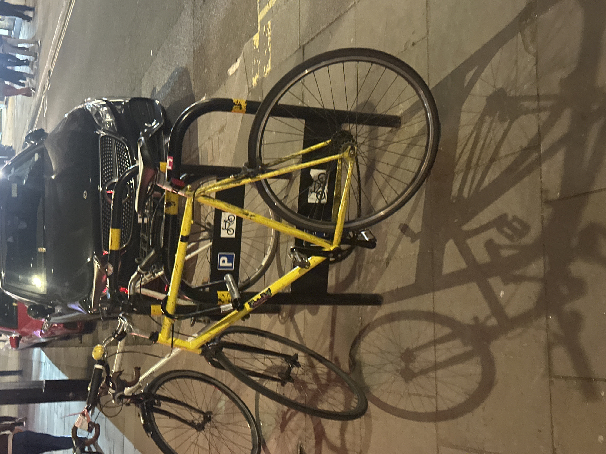 The image depicts an urban street scene at night or late evening, likely in London, with artificial lighting creating strong shadows. The main subjects are several bicycles, a black car, and people in the background.
In the foreground and central to the image, a bright yellow bicycle is prominently parked and secured to a black U-shaped bicycle rack. Its front wheel is turned slightly, and a visible sticker on its frame indicates "RALEIGH". Another white-framed bicycle is partially visible to the left of the yellow one, also seemingly secured. The strong light source casts long, detailed shadows of the bicycles and the rack onto the concrete pavement to the right.
To the left of the bicycle rack, a black car is parked. It appears to be a Mercedes-Benz, identifiable by the grille design and the three-pointed star emblem visible on the front. Its headlights are illuminated, reflecting off the shiny surface.
Further to the left, along what appears to be a sidewalk or curb, several people are visible, mostly from the waist down, suggesting they are walking. Their forms are somewhat blurred due to motion or low light conditions. Another person is partially visible in the bottom-left corner near a bicycle.
Notable details include distinct yellow double lines painted on the road or curb, characteristic of parking restrictions in the UK. On the black bicycle rack, a blue circular sign with a white "P" (for parking) and a small bicycle symbol are visible. The overall mood is quiet and typical of a city street after dark.