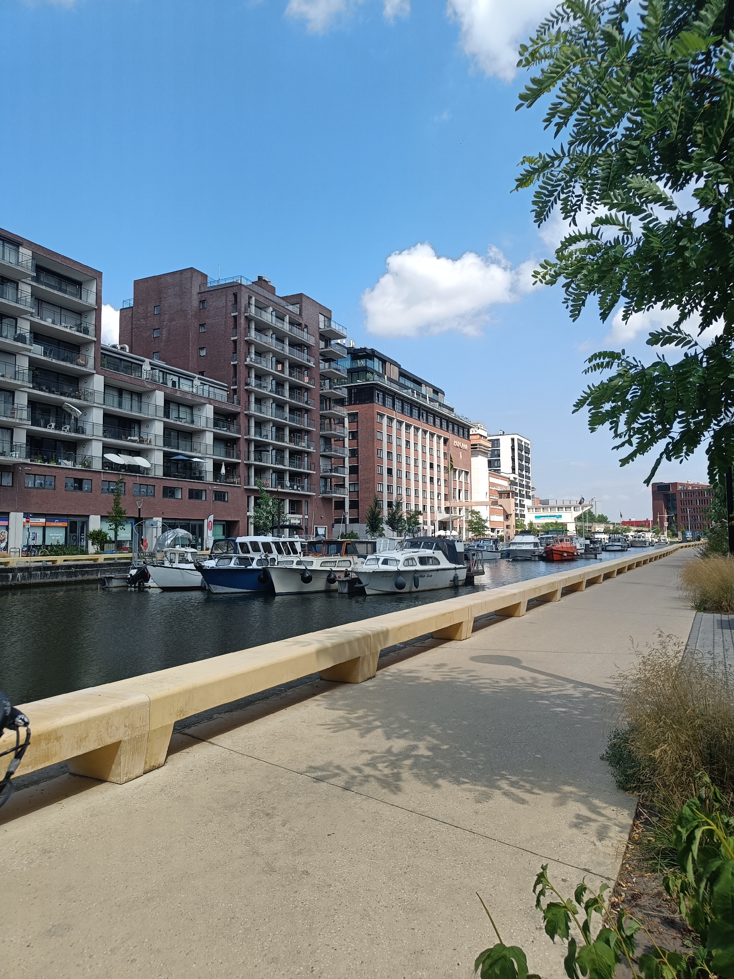 The image depicts a waterfront scene in a city, with modern apartment buildings lining the canal. Several boats are docked along the water, and a pedestrian walkway runs alongside the canal, bordered by a low concrete barrier. Trees and greenery are visible, with dappled sunlight casting shadows on the walkway. The sky is blue with scattered white clouds. The scene suggests a tranquil urban environment, possibly a residential or commercial district with recreational access to the water.