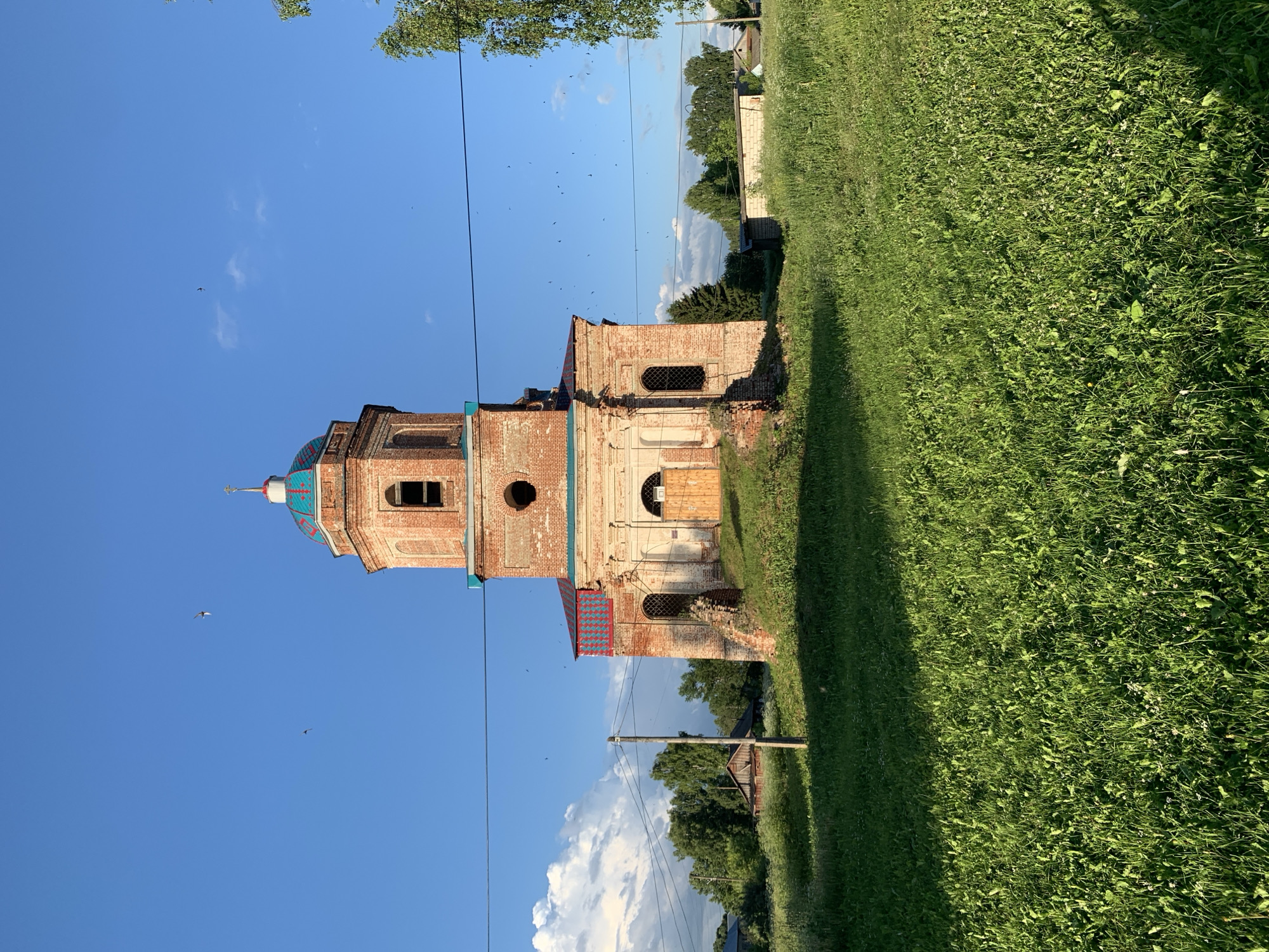 This is a full-frame, eye-level, outdoor photograph of an old, red brick church in the village of Svyatitsa, Russia, on a clear, sunny day. The church is the central subject, with its octagonal bell tower topped by a teal-colored dome with a cross. The church has arched windows and a wooden door. The building appears aged, with some signs of disrepair, particularly at the base where some bricks are missing or crumbling. In the foreground, there is a large expanse of green grass, with a shadow cast by an unseen object to the left. In the background, there are trees, some wooden buildings, and a power line stretching across the frame. The sky is a vibrant blue with a few scattered white clouds and numerous small, dark birds flying.