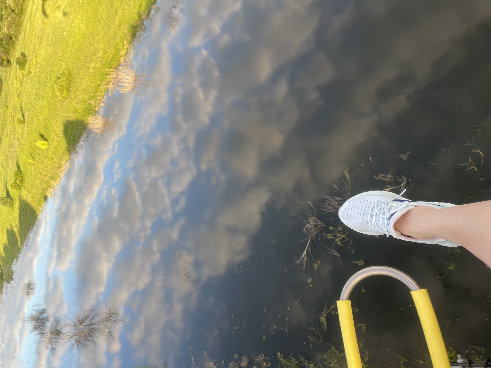 The image shows a first-person perspective of a person's leg and foot wearing a white sneaker, positioned next to a yellow handrail. The scene appears to be over a body of dark water, reflecting the sky with white clouds and a green grassy embankment with trees. This reflection suggests the vantage point is elevated, possibly from a hot air balloon or a bridge. The location is identified as Турово, Russia. The environment is natural, with water and vegetation. There are no other people clearly visible.