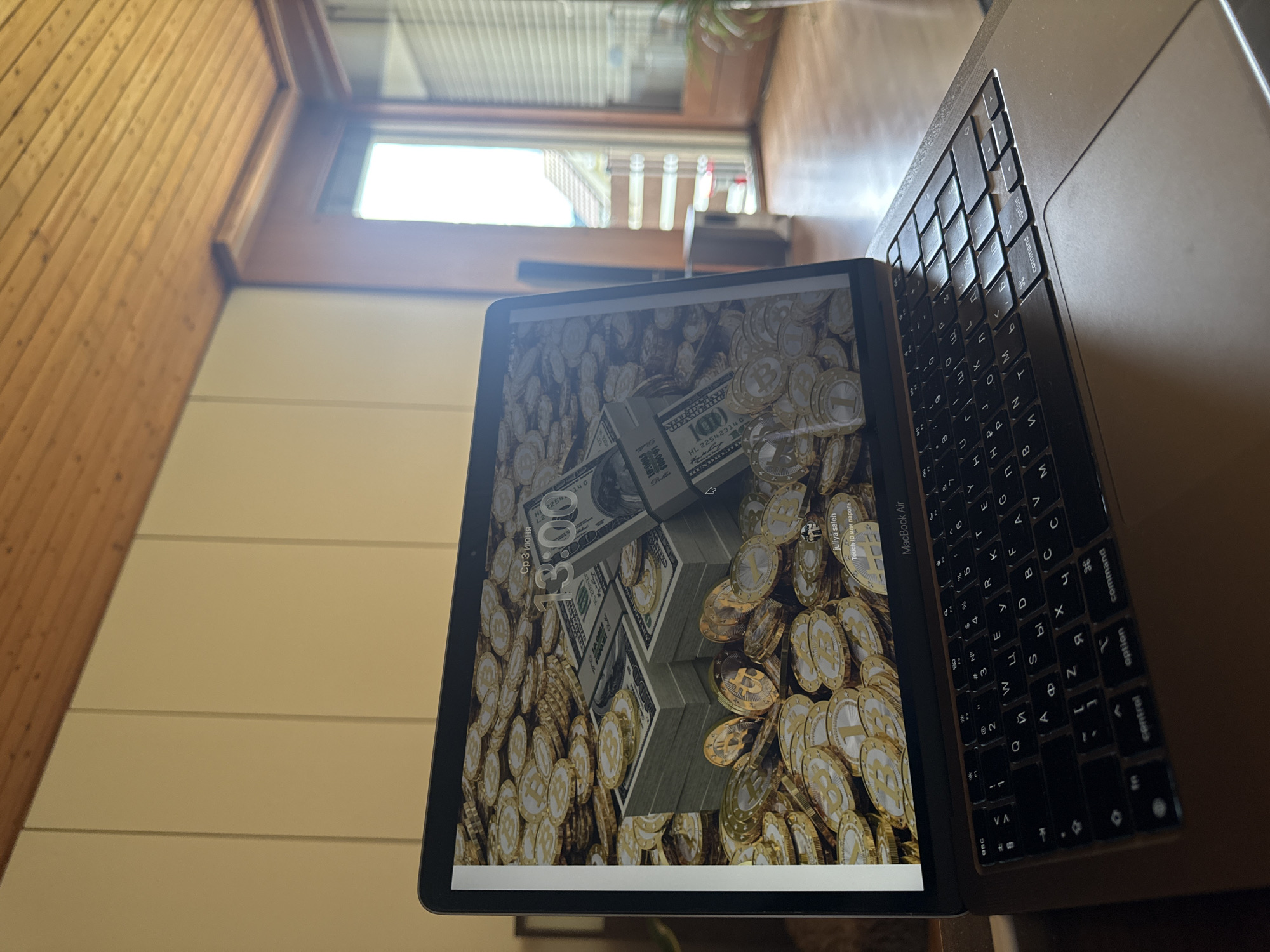 The image displays a close-up, top-down view of an indoor setting in Oberflachs, Switzerland, featuring a laptop and a tablet. A silver/grey MacBook Air laptop is partially visible on the right, with its black keyboard showing both Latin and Cyrillic characters. The tablet is positioned in front of the laptop, its screen prominently displaying an image of stacks of US dollar bills (hundreds) and numerous gold-colored coins, some featuring a 'B' symbol, indicative of cryptocurrency like Bitcoin.
The tablet's screen shows a lock screen or wallpaper with visible text: "Ср 3 июня" (Wednesday, June 3), "juliya saleh", and "Touch ID или пароль" (Touch ID or password). The dollar bills on the screen have details like "100" and "HL 22542314 G". The laptop's model name, "MacBook Air", is visible on its chassis.
In the background, a light-colored wall with horizontal lines is visible on the lower left, adjacent to a wooden slatted wall on the far left. A large window in the upper background allows natural light into the room, revealing a blurred view of an outdoor building structure or roof. Some indistinct greenery is also visible through the window. The overall scene suggests a personal workspace or a casual setup during daytime.