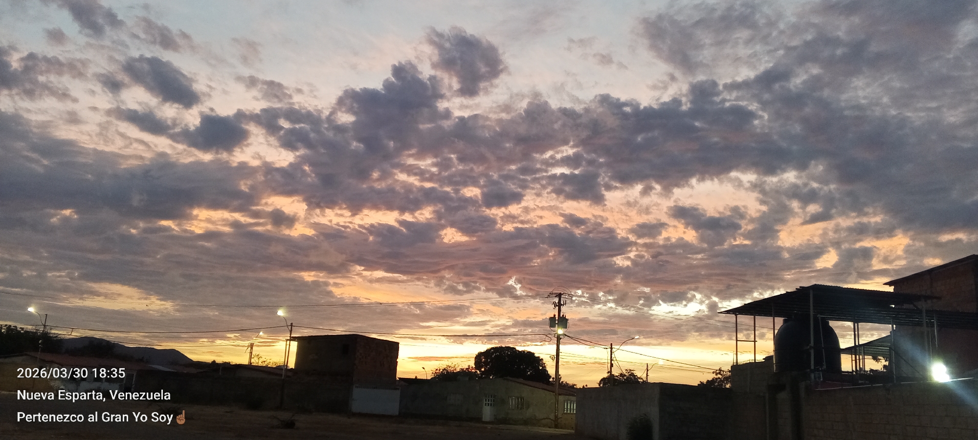 The image captures a twilight scene in Nueva Esparta, Venezuela. The sky is filled with striated cloud formations, tinted with hues of orange and grey from the setting sun. A series of low-lying buildings and structures populate the foreground. These include a brick building, houses, and an elevated structure with water tanks visible. A utility pole stands amidst the buildings with visible power lines. Distant hills are barely visible on the left horizon. The date "2026/03/30 18:35" is imprinted on the bottom left, along with the location, "Nueva Esparta, Venezuela." The text "Pertenezco al Gran Yo Soy" is also present.