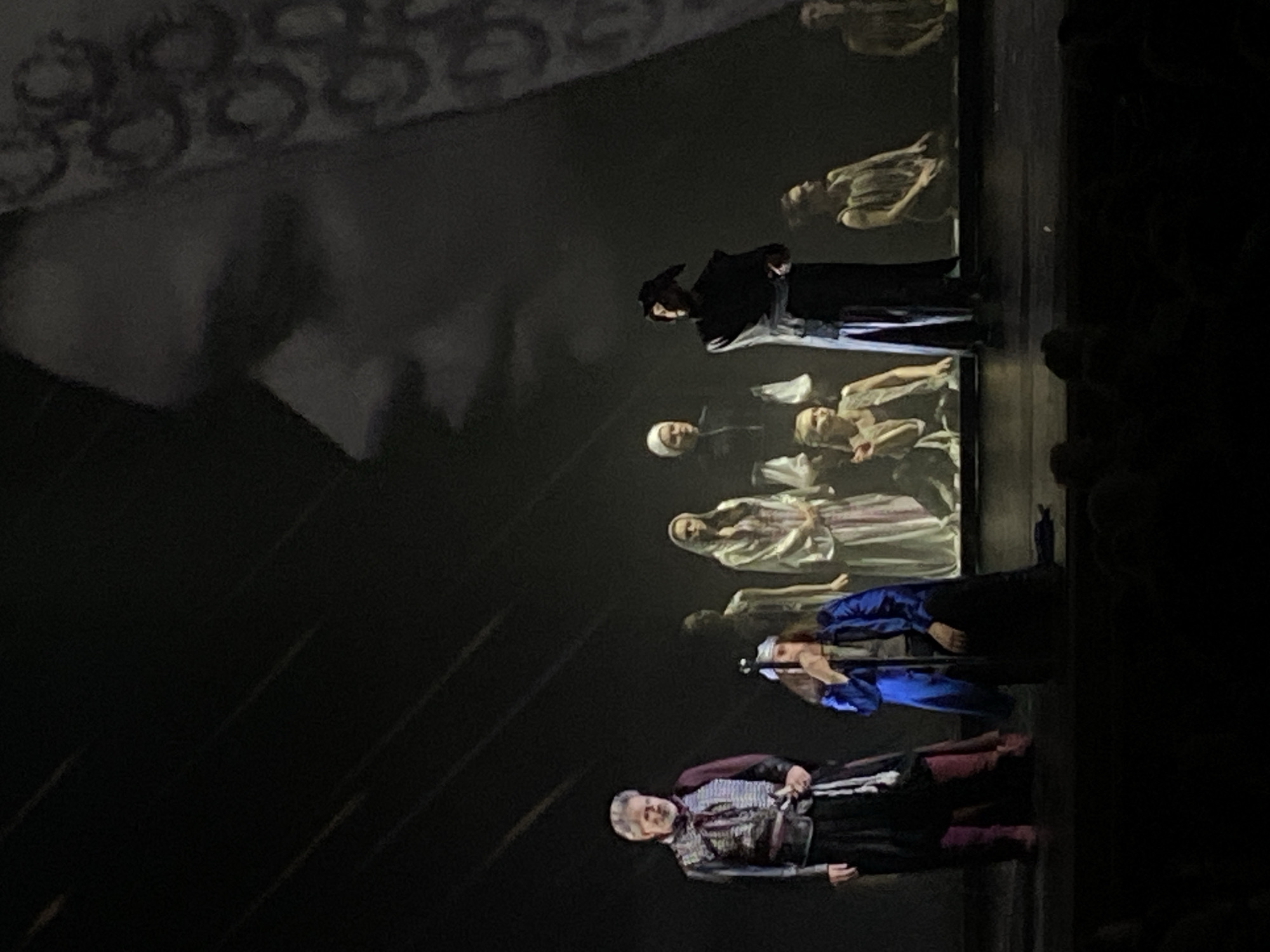 The image captures a scene from a theatrical performance, potentially a play or opera. Multiple actors are visible on stage, dressed in what appears to be historical or period costumes. A background projection or set design is present. The scene is dimly lit, creating a dramatic atmosphere. The location is likely a theater, though the city is unknown. The actors seem to be engaged in a performance. There are audience members present in the right of the frame.