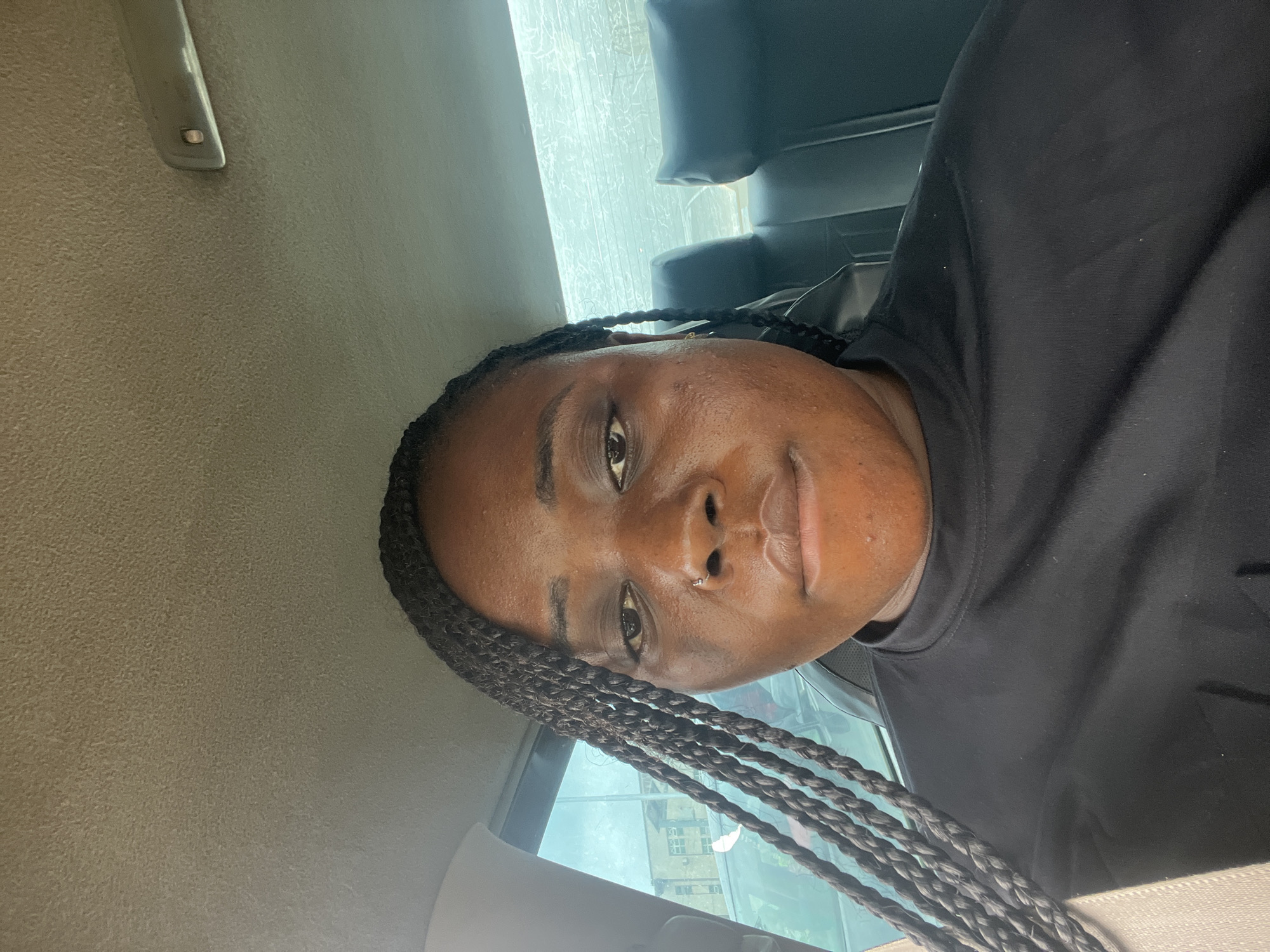 The image shows a woman inside a vehicle, possibly a car, with a light-colored ceiling and dark seats visible. She is wearing a black shirt and has braided hair. She appears to have a nose piercing. Through the car window, a building can be seen, indicating an urban environment. It is not possible to determine if the location is in the city of Abuja, Nigeria, or to know any more details.