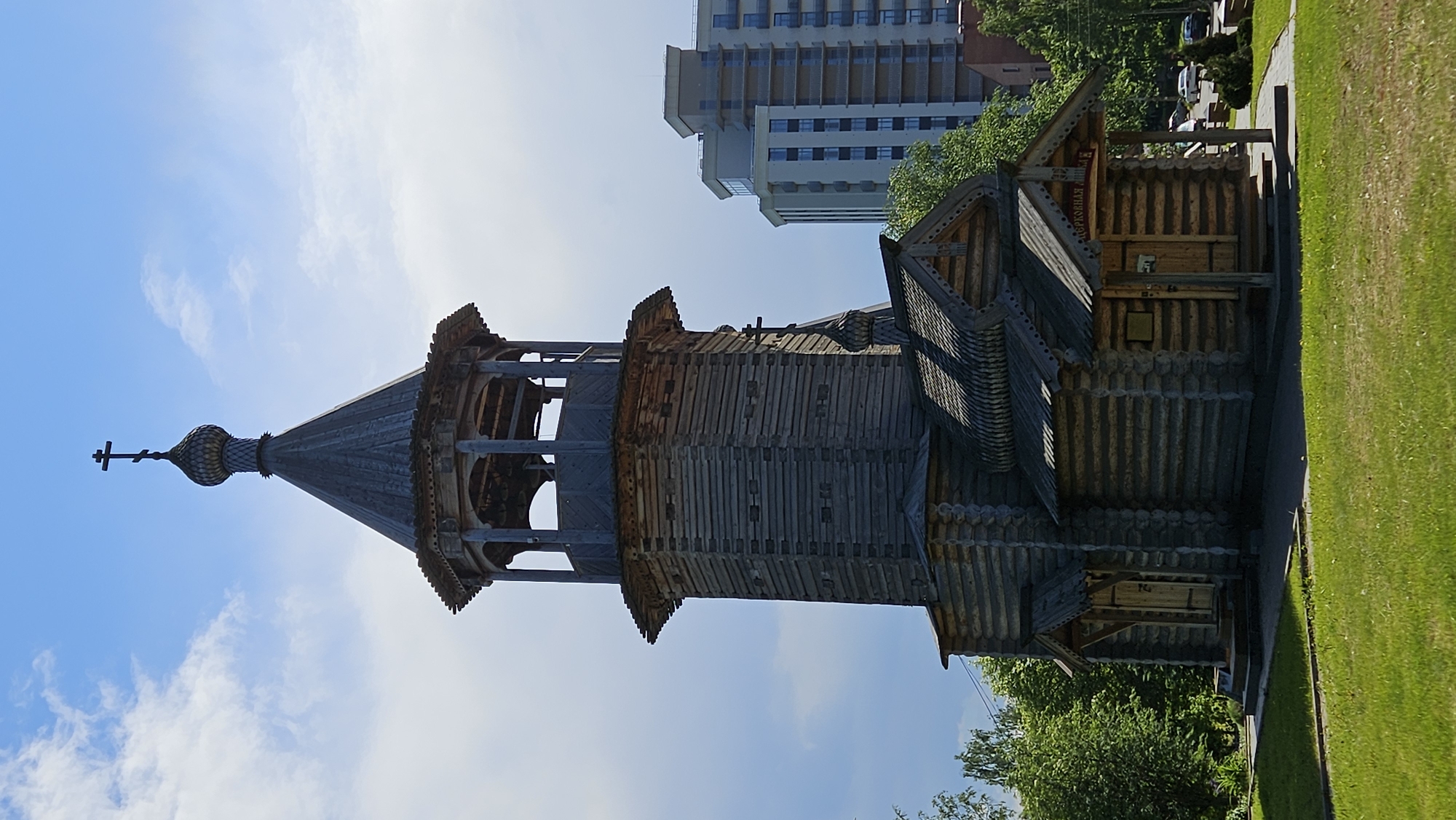 The image displays a traditional Russian Orthodox wooden church and bell tower under a bright blue sky with scattered white clouds. The primary structure is a tall, octagonal wooden bell tower, constructed from horizontally stacked logs, featuring an open belfry section and topped with a conical roof, a dark onion dome, and a Latin cross. Adjacent to the bell tower, a lower, smaller wooden building, likely the main church structure or an auxiliary building, also built from logs, has a gabled roof adorned with elaborate wooden shingles and a smaller onion dome topped with a cross.
Two wooden entrances are visible on the smaller building. The right entrance has a small gabled porch above it, bearing a red sign with white Cyrillic text that reads "ЦЕРКОВНАЯ ЛАВКА" (Church Shop). A dark plaque is affixed to the log wall to the left of this entrance. The immediate foreground consists of a well-maintained green grass lawn and a dark paved walkway leading towards the buildings. To the left, lush green trees are visible. In the background, to the right of the wooden complex, a modern, light-colored, multi-story building with numerous balconies stands, creating a stark architectural contrast. The scene is illuminated by bright sunlight.