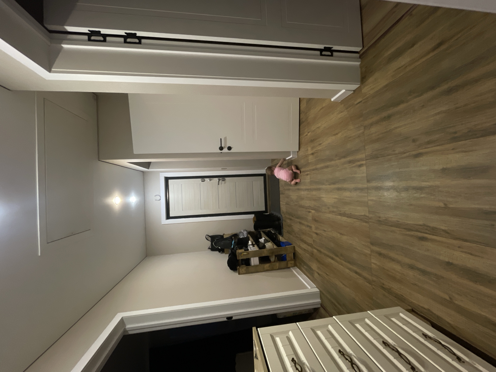 An interior shot captures a modern hallway or entryway, likely within a home, situated in Podshivalovo, Russia. The room features light-colored wooden flooring laid horizontally, contrasting with the crisp white walls and white trim. Artificial lighting, including recessed lights on the ceiling, illuminates the space.
In the foreground, towards the bottom right, part of a white cabinet or dresser with paneled doors and ornate dark handles is visible. Further into the hallway, a baby or toddler, dressed in a pink outfit, is crawling on the wooden floor, heading towards the left.
Along the left wall of the hallway, there are tall, white bi-fold doors with black bar handles, likely belonging to a closet. Further down this wall, another white built-in cabinet or door unit with small black knobs is present. At the far end of the hallway, a door with a black frame and multiple panels leads to another area.
On the right side of the hallway, near the far end, a light-colored wooden crate overflowing with various items, including shoes and bags, rests on the floor. A black bag is also nearby. Above this, a framed opening or window with white horizontal slats and a black frame is visible. A dark, horseshoe-shaped object is affixed to the wall next to this opening. An access panel is also visible on the upper left wall or ceiling. The overall impression is a well-maintained and functional domestic space.
