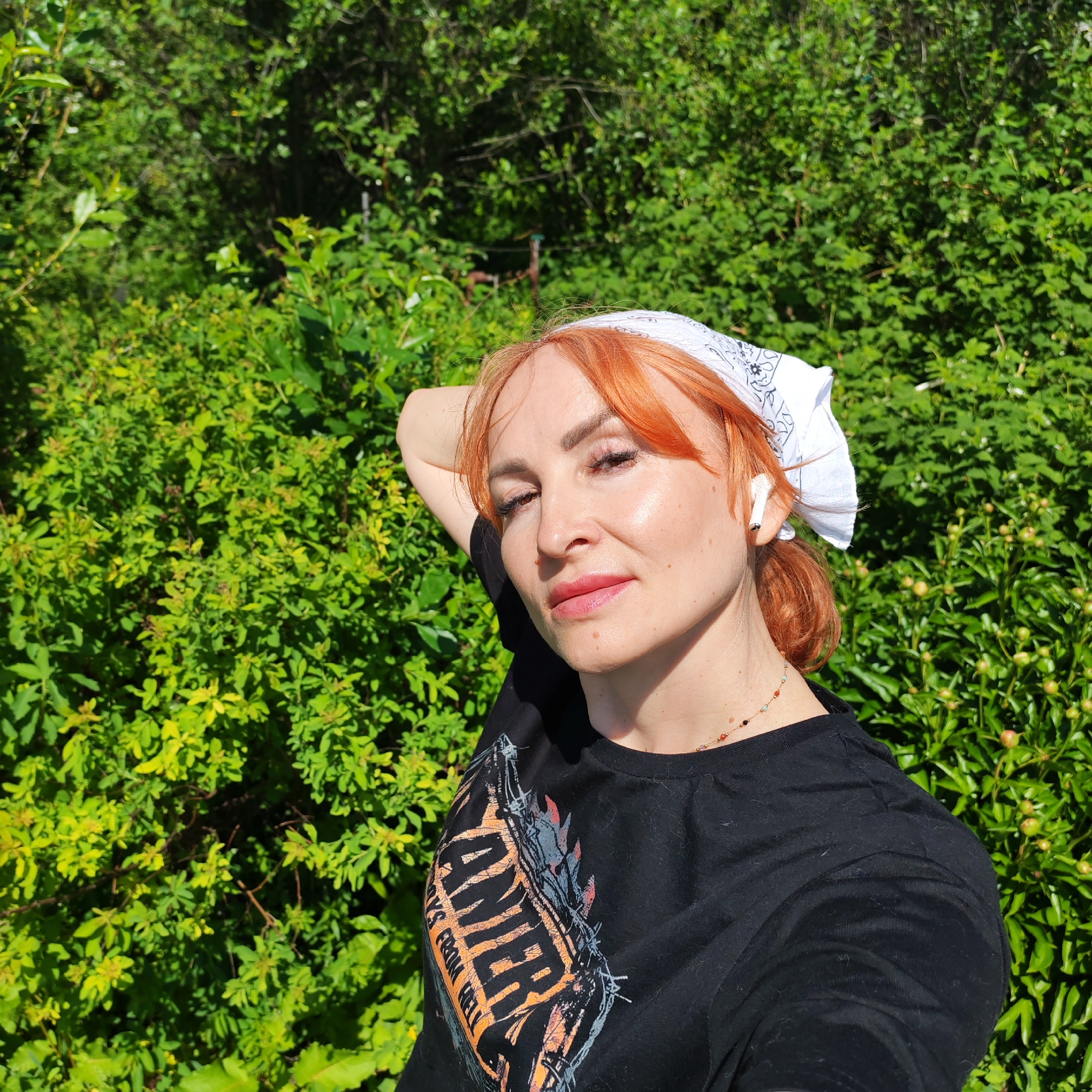 A woman with reddish-orange hair and fair skin is posing outdoors against a backdrop of lush green bushes. She wears a white bandana on her head and a black t-shirt with a graphic design. An AirPod is visible in her right ear. Her right arm is raised and resting behind her head. She has a necklace with small, colored beads. The location is Chaikovsky, Russia.