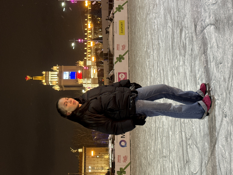 A woman in a black puffer jacket and blue jeans lies on an ice rink at night. She is wearing pink ice skates and faces the viewer. In the background, a brightly lit building with a red star and Russian flag colors can be seen. Various advertisements and logos border the rink. The ice surface displays skate marks. It appears to be a winter scene.