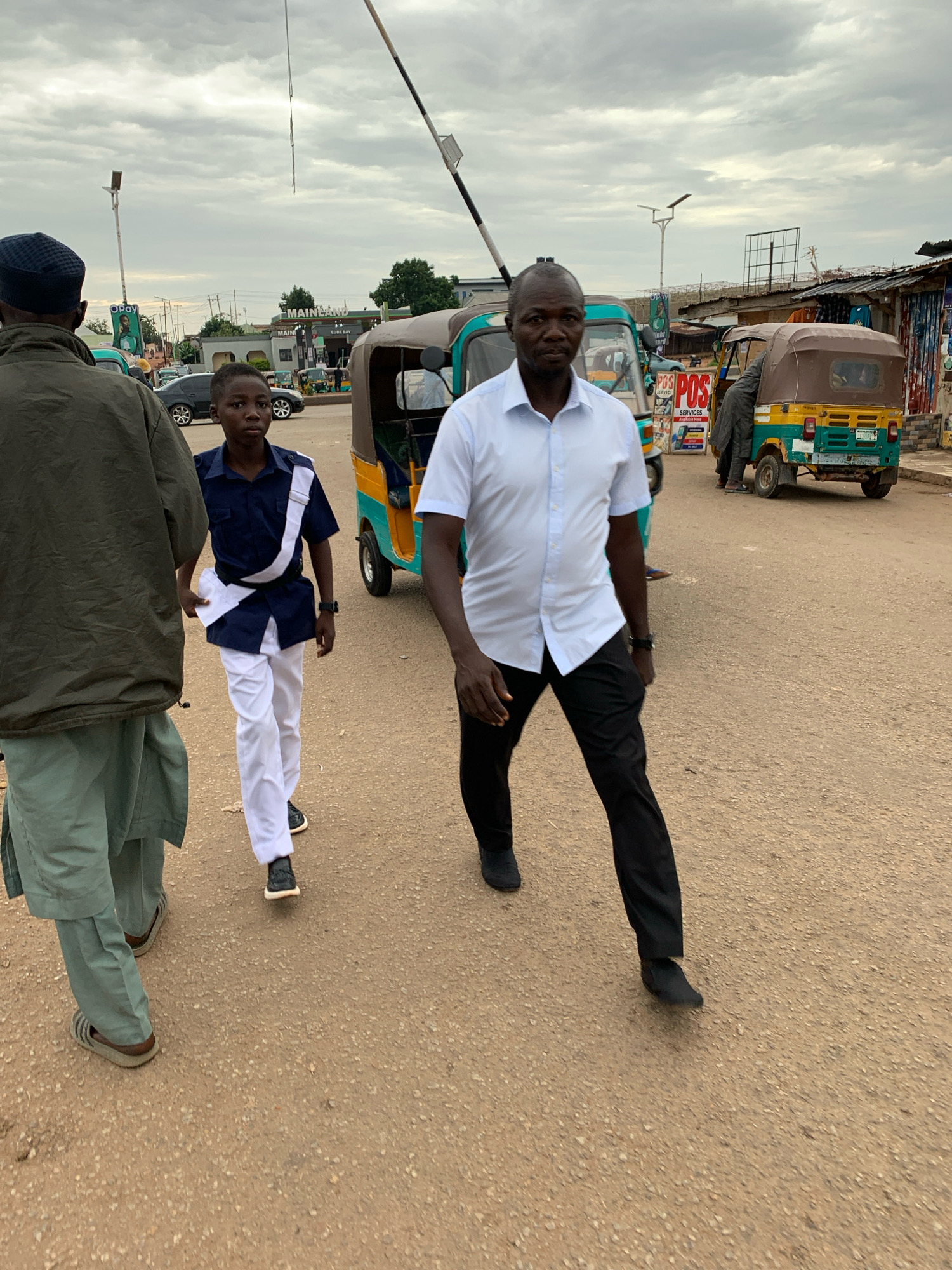 In Gura Top, Nigeria, under an overcast sky, three individuals walk on a dusty road. The person on the left, seen from behind, wears a dark cap, a green jacket, and light-colored trousers with sandals. A young boy in the center, facing forward, is dressed in a dark blue uniform with a white sash and white trousers, black shoes, and appears to be walking alongside the man. The man on the right, in focus and walking towards the camera, wears a light blue collared shirt, black trousers, and black shoes. Behind them, two auto-rickshaws, one yellow and green and the other predominantly green, are parked or moving on the road. In the background, various shops with signs, including "POS SERVICES," and structures line the street, and utility poles are visible against the cloudy sky. A retractable barrier is also present, suggesting a checkpoint or entrance.
