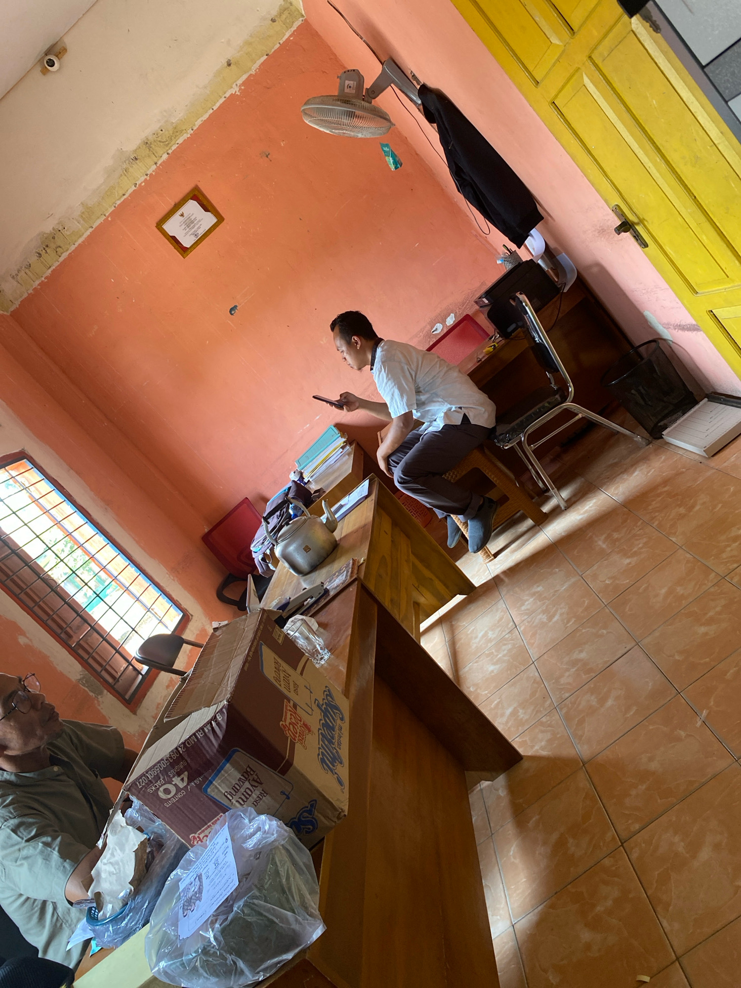 The image depicts an indoor scene, possibly an office or a small shop, with an orange-painted wall and a yellow-painted door visible. The floor is tiled with terracotta-colored squares. A man in a light-colored shirt and dark pants is seated at a wooden desk, holding a mobile phone. Another man, wearing glasses and a greenish shirt, is seen in the foreground, looking at a large cardboard box. There are other objects in the room, including a ceiling fan, a security camera, a framed certificate on the wall, a kettle, and stacks of boxes and plastic-wrapped items on the desk. The overall impression is of a functional, albeit somewhat cluttered, workspace. The location is stated to be in the city of Mojo Pahit, Indonesia.