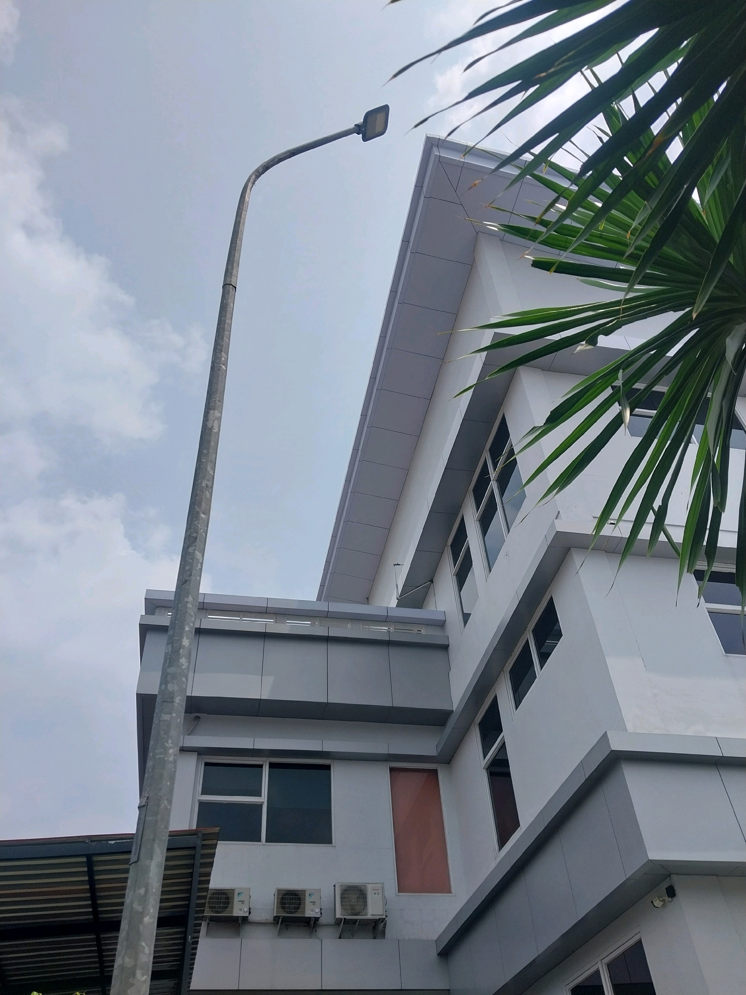 The image depicts a modern white and gray building with a blue sky and some clouds in the background. The building has a geometric design with multiple levels and several windows. A street lamp is positioned to the left of the building, and some palm fronds are visible in the upper right corner, suggesting a tropical or subtropical climate. Three air conditioning units are mounted on the lower section of the building. The scene appears to be an outdoor shot, possibly in an urban or commercial area, given the architecture and the street lamp. The lighting suggests it is daytime, possibly overcast due to the cloud cover. The image is taken from a low angle, looking upwards towards the building and sky. The location is identified as Sleman, Indonesia.