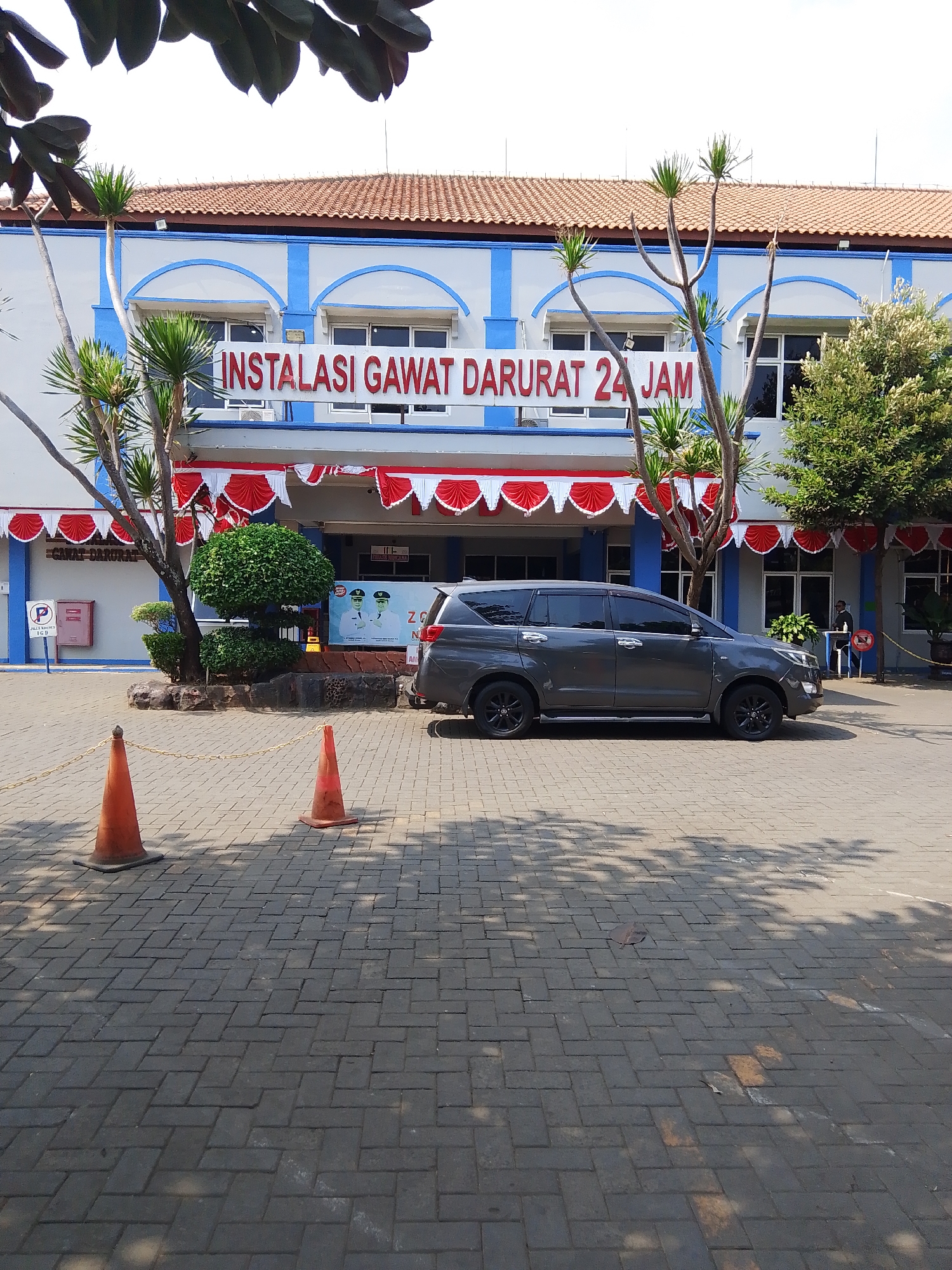 The image shows the entrance of a hospital's emergency room, labeled "INSTALASI GAWAT DARURAT 24 JAM". The building has a white facade with blue trim and a terracotta tiled roof. Red and white pennant decorations are hung above the entrance. In front of the building, there is a gray SUV parked on a brick paved area. Two orange traffic cones are placed on the pavement in the foreground. Trees and plants are visible around the building. A sign with a blue 'P' indicates a parking area. The scene appears to be in Potroyudan, Indonesia.