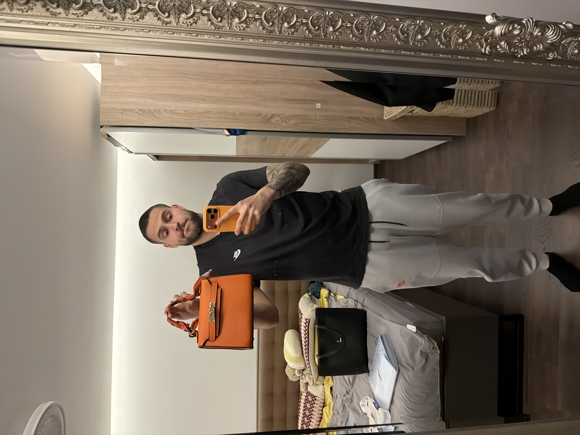A medium-shot image captures a man taking a mirror selfie. He stands in front of a large mirror framed with ornate silver detailing. The man has a beard and is wearing a black t-shirt and light grey sweatpants. He holds an orange handbag in one hand and an orange smartphone in the other. Reflected in the mirror behind him is a bedroom setting with a bed and other personal items. The room's color palette includes white, beige, and dark brown. The location is indicated to be Sofia, Bulgaria, but this is not visibly verifiable in the image.
