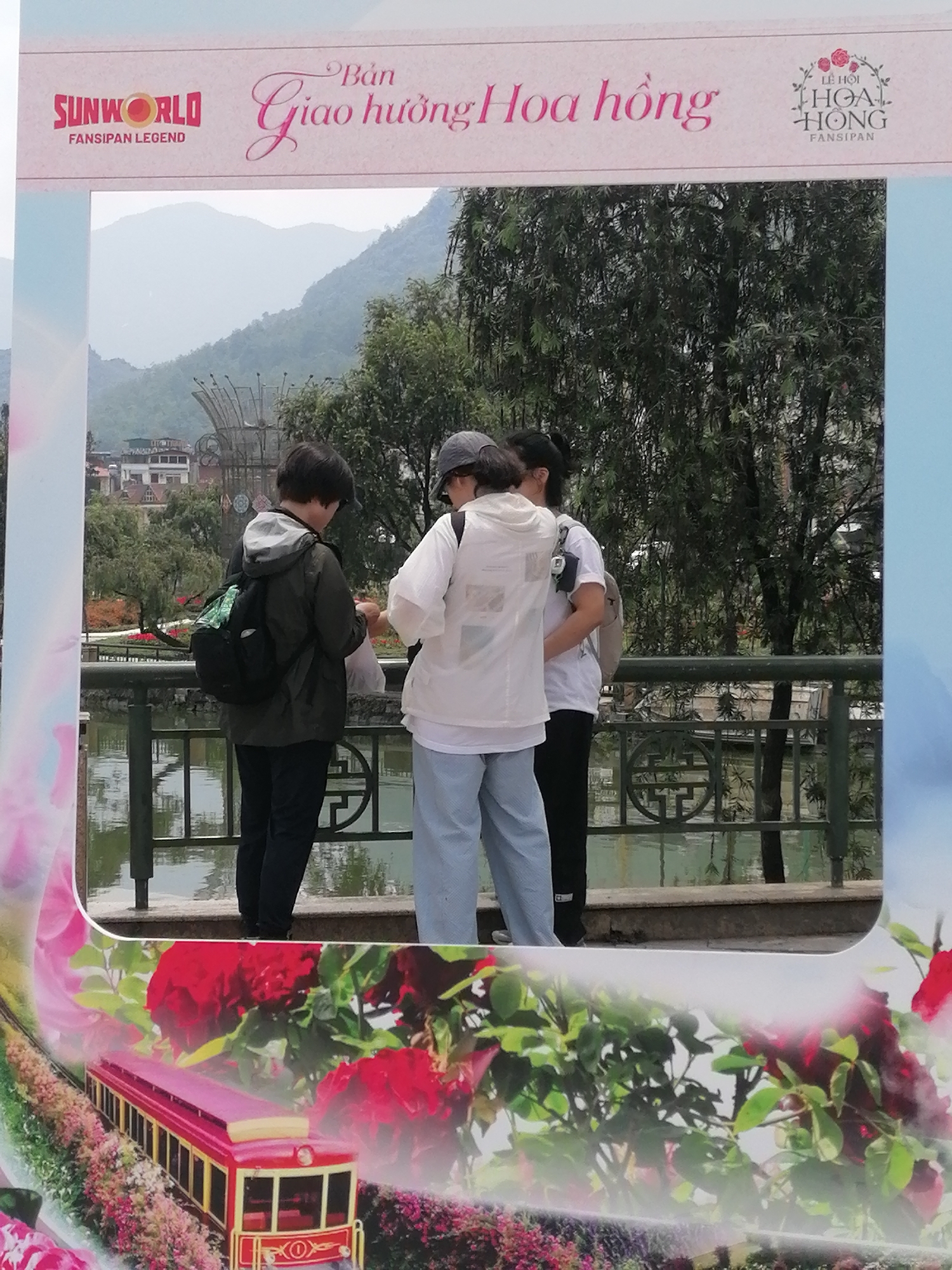The image shows three young adults standing by a bridge in the city of Sa Pa Ward, Vietnam. The person on the left is wearing a black jacket with a hood, black pants, and a backpack. The person in the middle is wearing a white shirt, light blue pants, and a cap. The person on the right is wearing a white shirt and black pants. They are standing behind a metal railing. The background shows trees, buildings, and mountains. The image is taken through a frame, with "SUNWORLD FANSIPAN LEGEND" and "LỄ HỘI HOA HỒNG FANSIPAN" written on the top border and an image of a red and gold train surrounded by flowers on the bottom border.