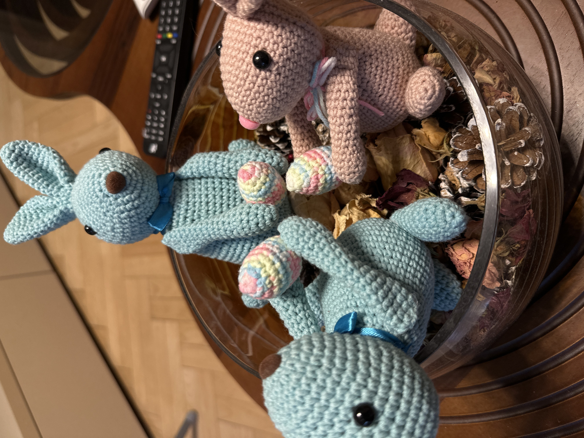 The image shows three crochet bunny toys, two blue and one pink, inside a glass bowl filled with dried leaves and pinecones. The bowl sits on a dark wooden surface. A television remote is visible in the background. The image was taken in Stara Zagora, Bulgaria.