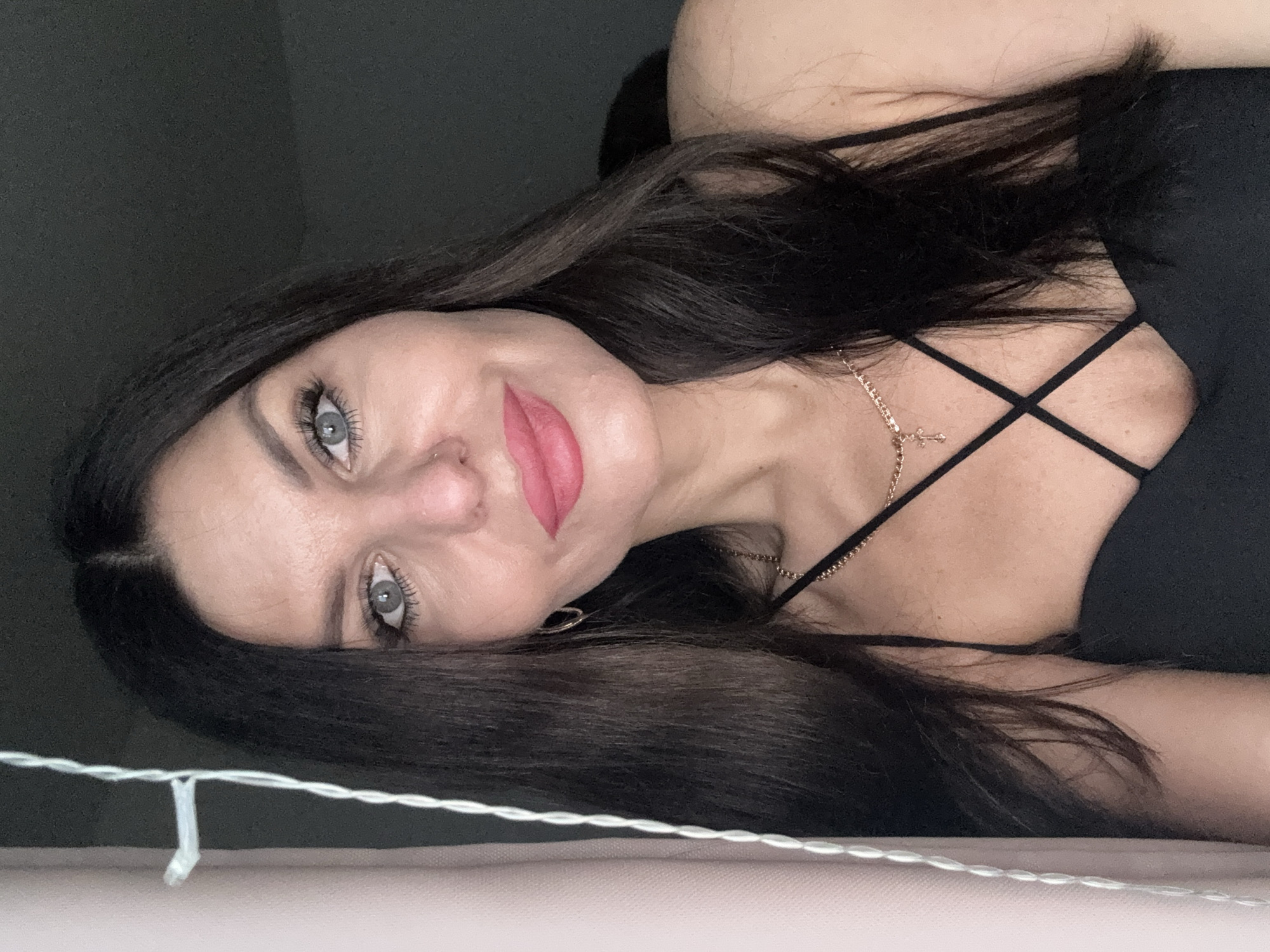 A close-up, waist-up portrait of a woman with dark hair and light blue eyes is featured in the image. She is wearing a black spaghetti strap top and a delicate gold necklace with a cross pendant. Her makeup includes pink lipstick and mascara. To the left of the frame, a portion of a pink curtain and a white string of lights are visible, suggesting an indoor setting, possibly a home. The background is dark and out of focus. The image does not provide any information or visual cues about the city of Chaikovsky, Russia.