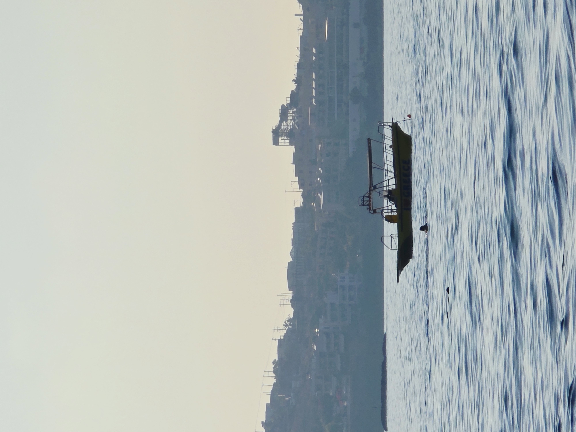 In Malia Municipal Unit, Greece, a yellow boat with a covered top is on the water. The boat has the word "POLICE" written on its side. Two people are visible on the boat, one standing and one sitting. In the water near the boat, two individuals are swimming. In the background, a hillside is covered with buildings and scattered trees, with power lines visible among them. A large structure that appears to be a water slide is also visible on the hillside. The sky is a pale, hazy yellow.