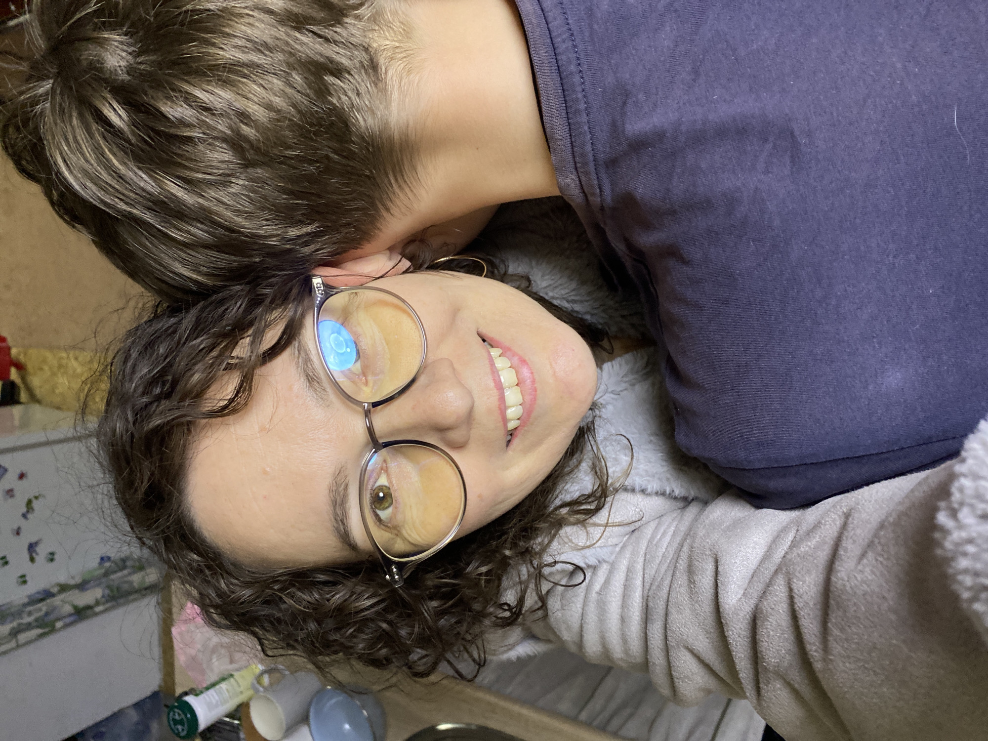 A selfie shows a woman with curly dark brown hair and glasses smiling. A person with short brown hair is nuzzling her head/neck. The woman is wearing a light-colored fleece jacket. The other person is wearing a dark purple t-shirt. In the background, there are items on a counter including containers, cups, and a steel bowl. There is no evidence to indicate a location in the city of Noviy, Russia.