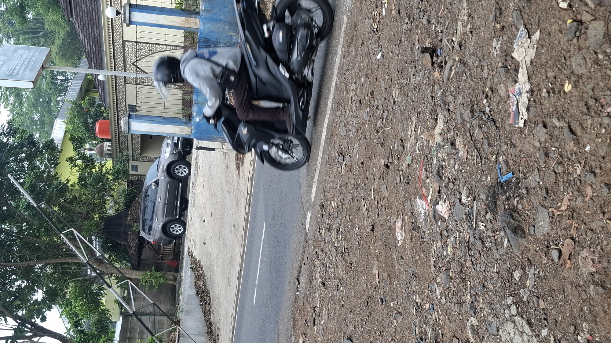 The image depicts a street scene in Cilaku, Indonesia. In the foreground, a person is riding a black scooter, wearing a grey hoodie and a black helmet. The scooter is moving on a paved road with a white line marking the lane. To the left of the road, there is a rough, unpaved area with debris, including stones, dirt, and scattered litter. In the background, a brown SUV is parked on a slightly elevated area. Behind the SUV, there is a building with a green facade and a dome, possibly a mosque. A red water tank is visible on top of the building. A sign with Arabic script and text in Latin characters is mounted on a pole near the building, indicating "MASJID BABUSSALAM". A blue barrier runs along the edge of the road in the middle ground. The environment appears to be a mix of urban and natural elements, with trees and foliage visible in the background. The lighting suggests it is daytime.