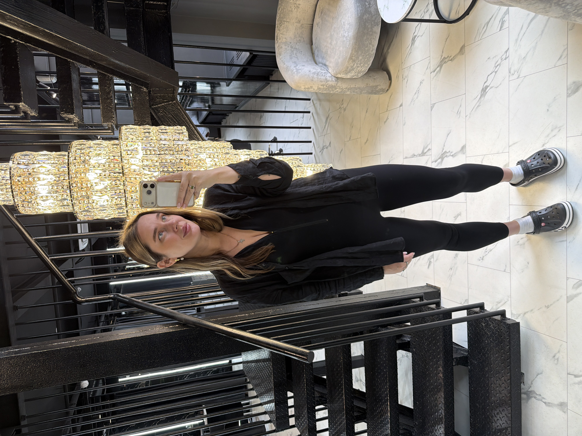 A young woman is captured taking a mirror selfie within a modern, brightly lit interior, likely in Moscow, Russia. She is reflected in a large, multi-tiered crystal chandelier that occupies the left side of the frame, emitting a warm glow. The woman has light brown hair and is dressed in a black long-sleeved top, black leggings, and black Crocs shoes paired with white socks. She holds a light-colored smartphone, possibly an iPhone, up to capture her reflection. Her face is visible, looking slightly upwards with a neutral expression, and she wears earrings, a necklace with a small cross, and a ring.
The setting features a floor composed of white tiles with a subtle grey marble-like pattern. To the right, a plush, light-colored upholstered armchair and a small round white table are visible. A prominent black metal staircase with a sturdy railing and textured steps runs through the lower and left parts of the image, suggesting an industrial-chic or contemporary design. Additional black architectural elements are present in the background. The overall scene conveys an upscale commercial or residential environment. The lighting suggests it is daytime or a well-illuminated indoor space. No visible text is present in the image.