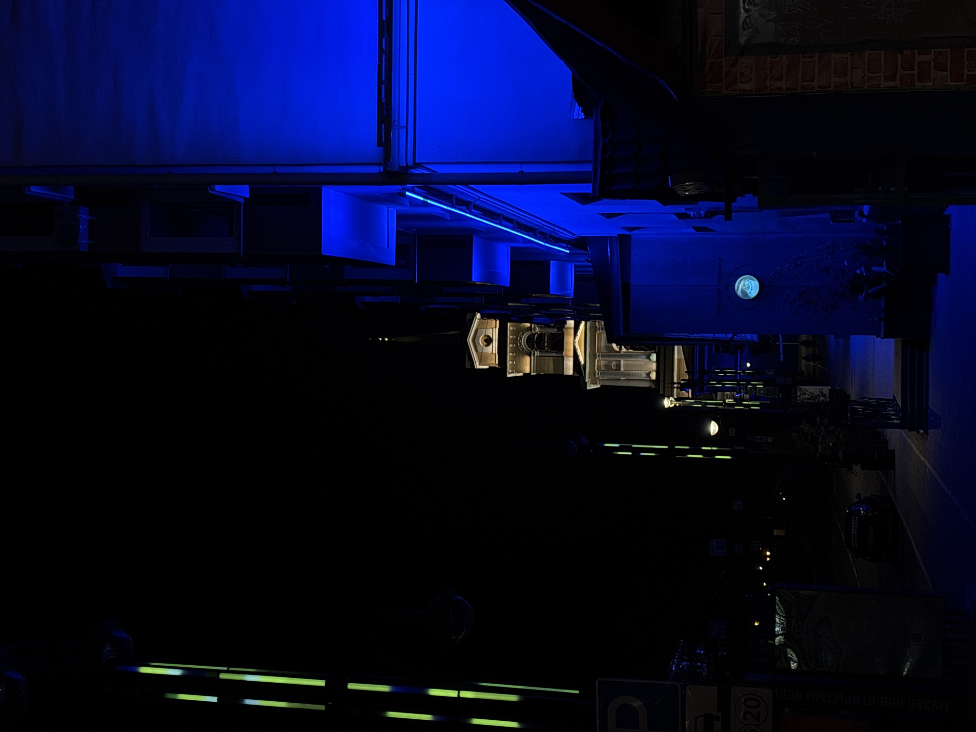 The image depicts a street scene in Perm, Russia, at night. The photograph is taken from a ground-level perspective. Illuminated buildings and streetlights cast light on the pavement, with the lower portion of the frame primarily dark. A neon sign emitting green light is visible at the bottom of the frame. Overhead, structures are illuminated with blue light. The scene includes architectural elements and a glimpse into the layout of the street.