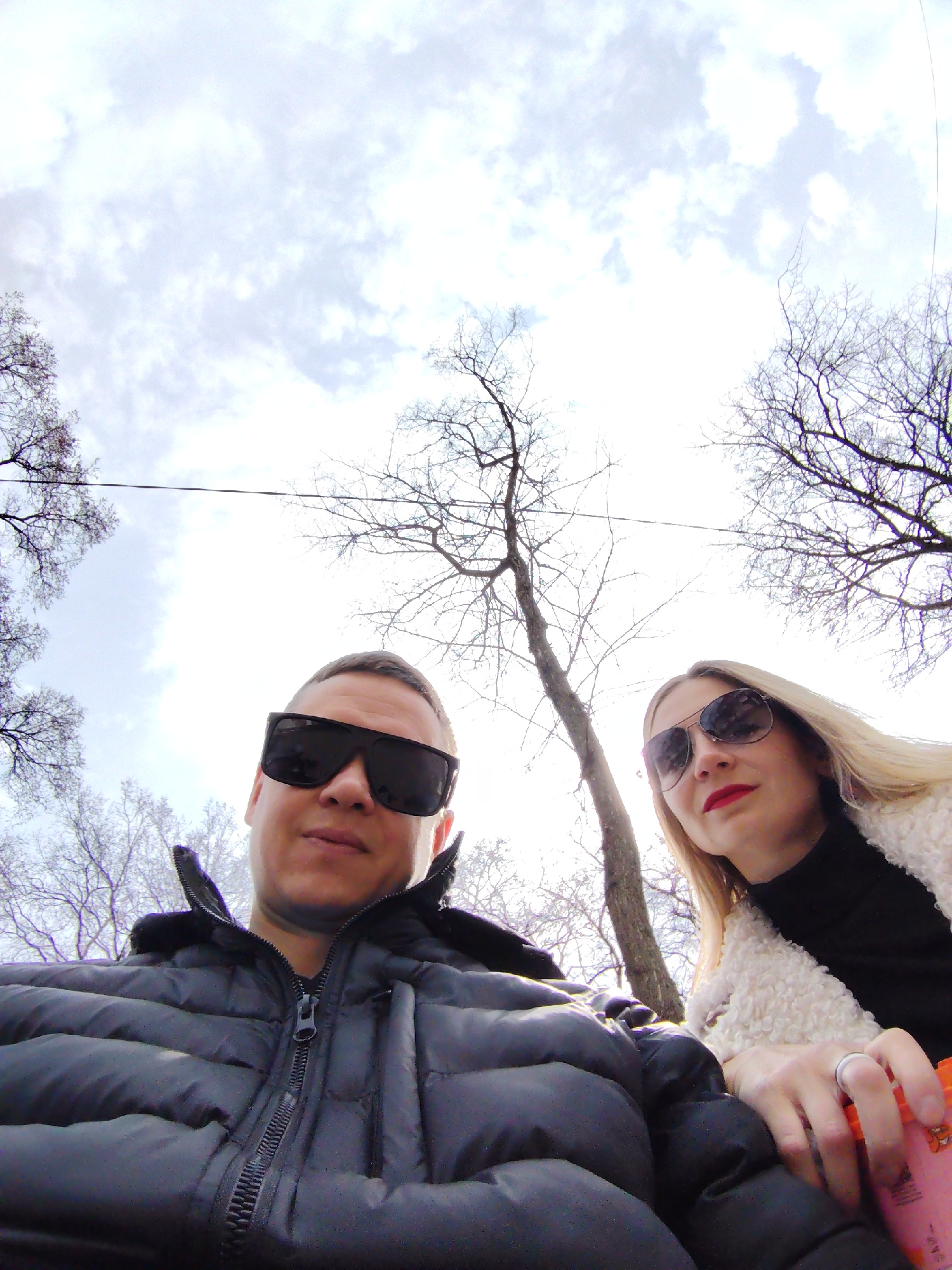 A selfie captures two individuals outdoors. A man with short, dark hair and dark sunglasses is wearing a black puffy jacket, and a woman with blonde hair and dark sunglasses is wearing a black turtleneck and a white sherpa jacket. They are positioned close together, with the camera angled upwards, capturing the sky and bare tree branches above. The background consists of a cloudy sky and trees with bare branches, a detail indicating it may be winter or early spring. In the woman's hand, there is a pink carton. The location is Chaikovsky, Russia, though specific geographical features aren't clearly definable.
