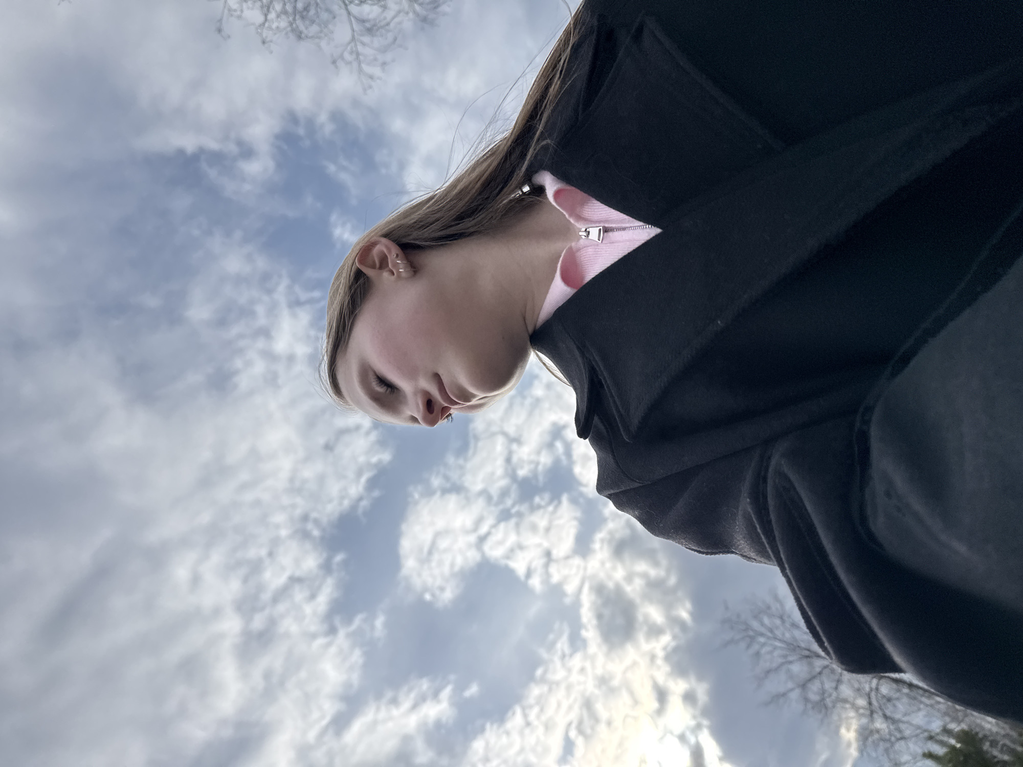 The image is captured from a low, upward-looking perspective, with the camera pointed towards the sky. A young person, likely female, is the primary subject, seen from a very low angle, looking downwards. Their face is partially visible in profile, showing their nose and chin, with their eyes appearing closed or cast downwards. They have fair skin and long, light brown hair, which falls over their right shoulder. A small hoop earring or piercing is visible on their right ear.
The individual is wearing a light pink, possibly zipped-up top, layered under a dark, presumably black, heavy jacket or coat, the texture of which suggests a wool-like material. The zipper pull on the pink top is silver.
The background is dominated by a cloudy sky, with white and grey clouds scattered across a lighter blue sky. The lighting suggests it is daytime, with soft, diffused sunlight. In the upper left corner, bare tree branches are silhouetted against the sky, indicating a winter or early spring season. Faint, darker foliage, possibly evergreen trees, can be seen in the lower right, partially obscured by the person's clothing. The scene depicts an outdoor setting, consistent with the provided location context of Chaikovsky, Russia. No visible text is present in the image.