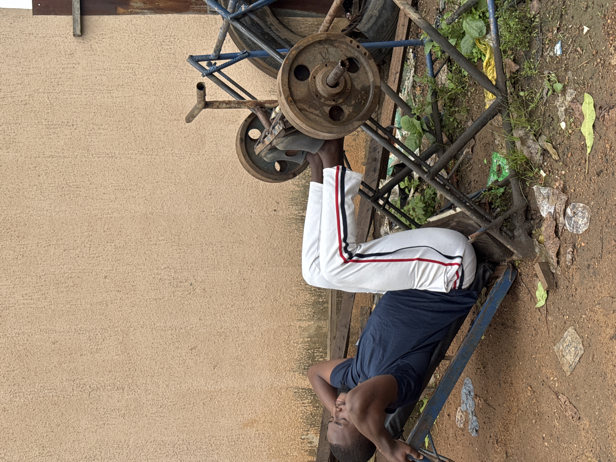 A young Black man is performing a leg press exercise on a makeshift weightlifting machine outdoors. He is lying on his back on a blue inclined bench with his hands behind his head, and his feet are on the weight plate. He is wearing a dark blue t-shirt and white athletic pants with red and black stripes down the sides. The weightlifting machine appears to be constructed from metal bars and has large, rusty weight plates. The ground is dirt, littered with debris such as plastic bags, leaves, and what looks like a piece of cardboard. In the background, there is a textured, light brown wall and some metal scaffolding with a large tire. The setting appears to be an outdoor gym or a yard in a residential area in Akwanga, Nigeria. The lighting suggests it is daytime.