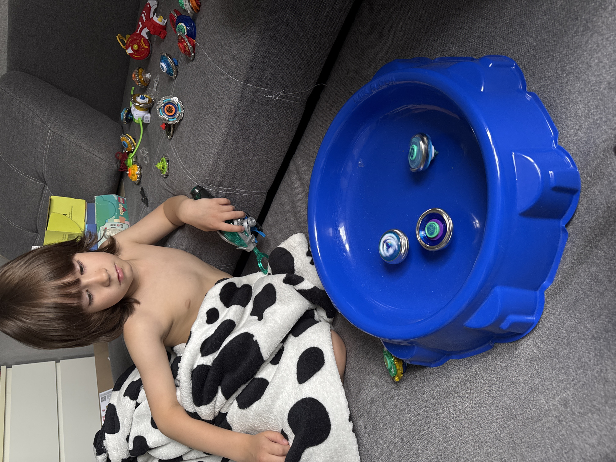 A young child with brown hair is lying on a gray couch covered with a black and white spotted blanket. The child is shirtless and holding a toy launcher in their right hand. A blue toy arena containing three spinning tops sits next to the child on the couch. Several more spinning tops and toy components are scattered on the couch behind the child. The scene appears to be indoors. There is no indication of the location being Chaikovsky, Russia, or any other city.