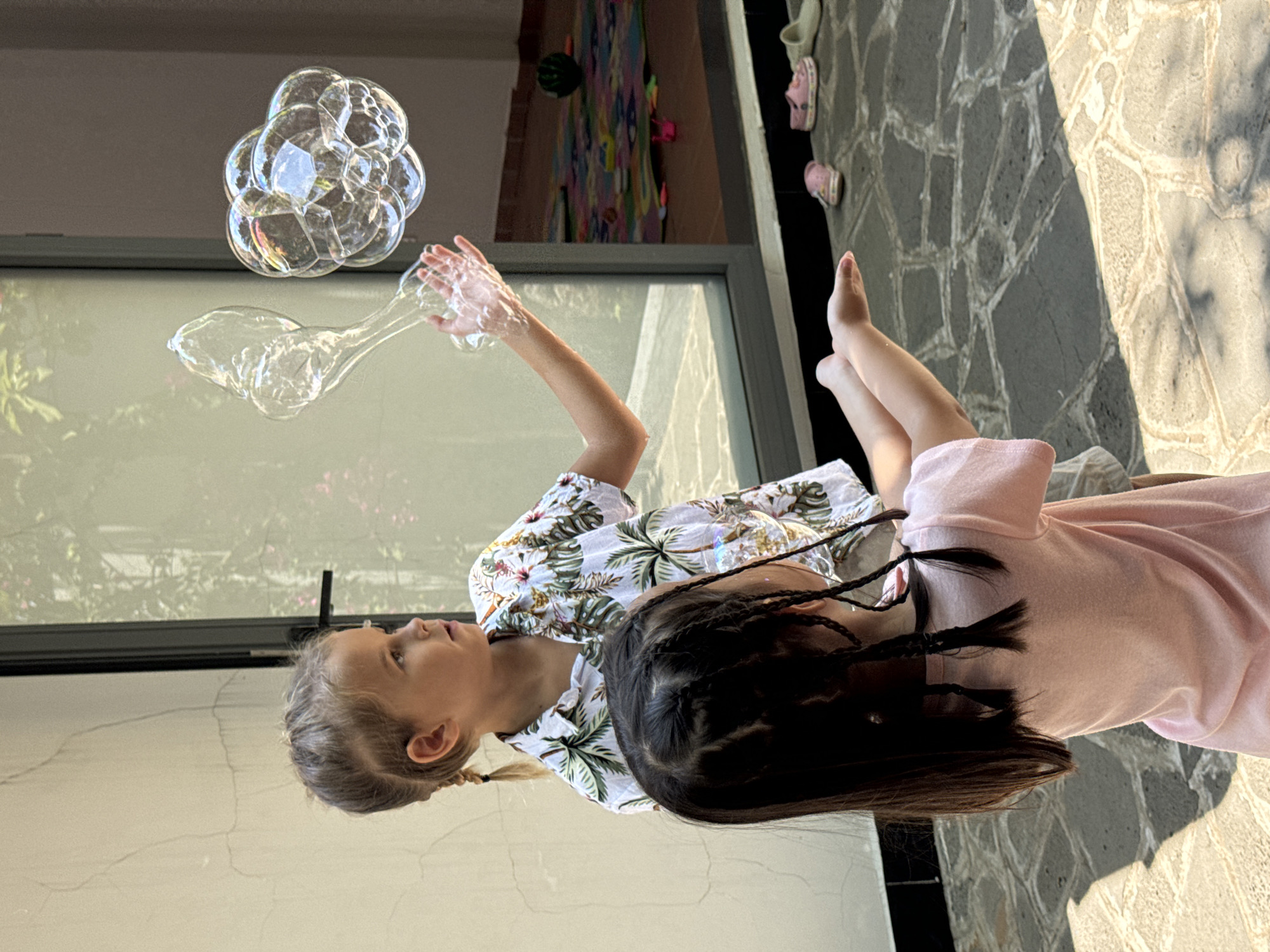 Two children, likely girls, are positioned outdoors. One, wearing a white, short-sleeved shirt with a tropical print, is reaching towards a cluster of soap bubbles, which are floating in mid-air. The other, wearing a pink dress, has dark hair styled in braids. The location appears to be outdoors, with a stone-paved area on which shadows fall. In the background, a partially visible window reveals a colorful mat or carpet. The location is not specified as Mũi Né, Vietnam in the photo.