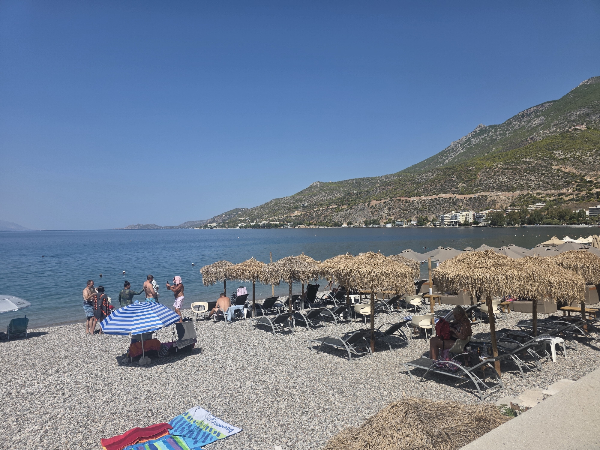 The image depicts a beach in the Municipal Unit of Loutraki - Perachora, Greece, on a clear, sunny day. The beach is covered in grey pebbles, and the sea is a calm, deep blue. In the foreground, several beach chairs and umbrellas are set up. Some people are visible on the beach. To the left, a group of people is gathered near the water, with one person taking a photo. Further to the right, several people are seated on lounge chairs under thatched umbrellas. In the background, a town with white buildings stretches along the coastline, nestled at the base of a lush green, mountainous landscape. The sky is a vibrant, cloudless blue.